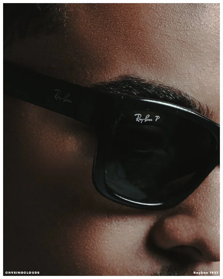 Close-up of a person's face wearing black Ray-Ban sunglasses, showing their eyebrow, partially visible nose, and dark skin tone.