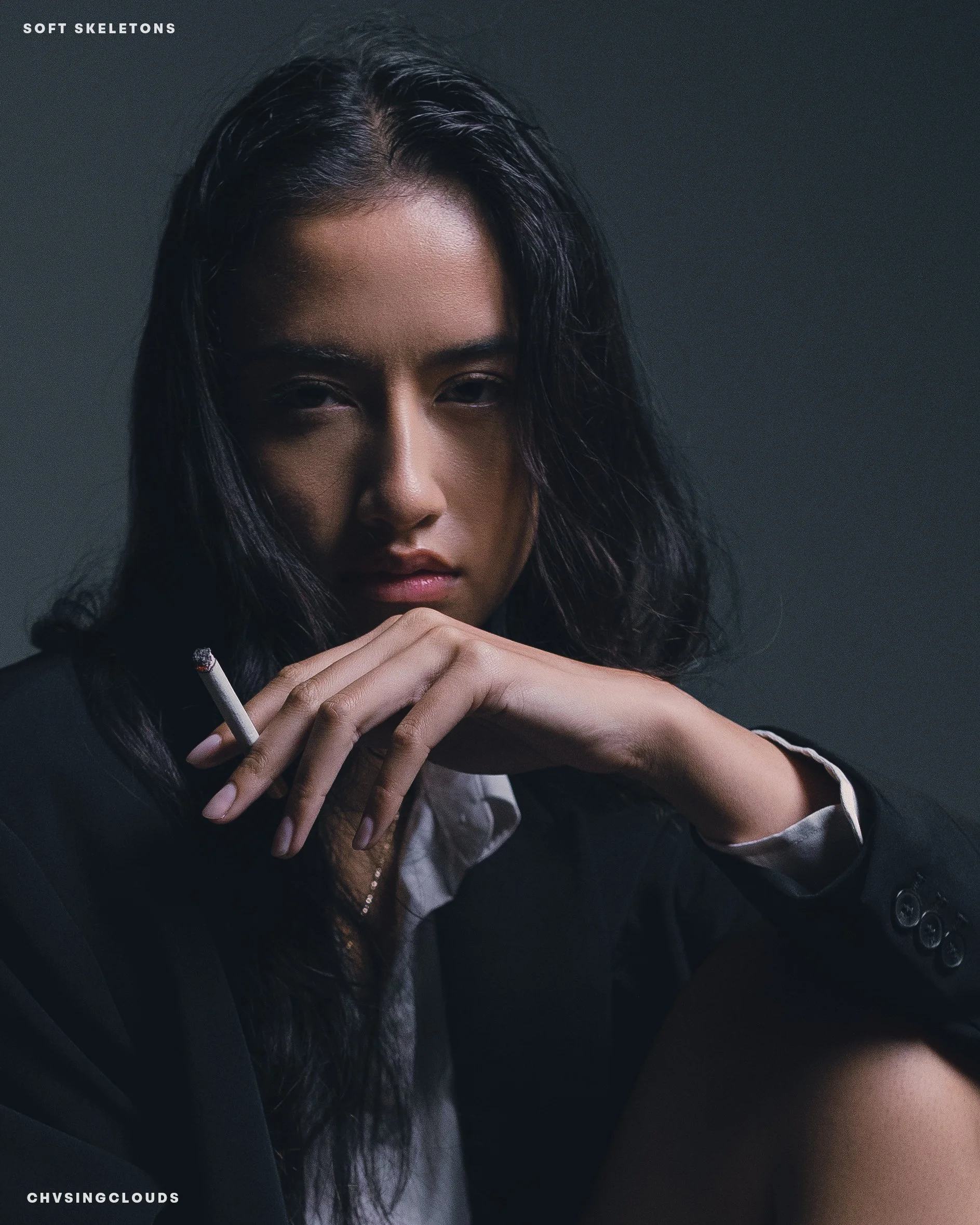 Portrait of a woman with dark, wavy hair holding a lit cigarette, wearing a black blazer and a white shirt, against a dark background.