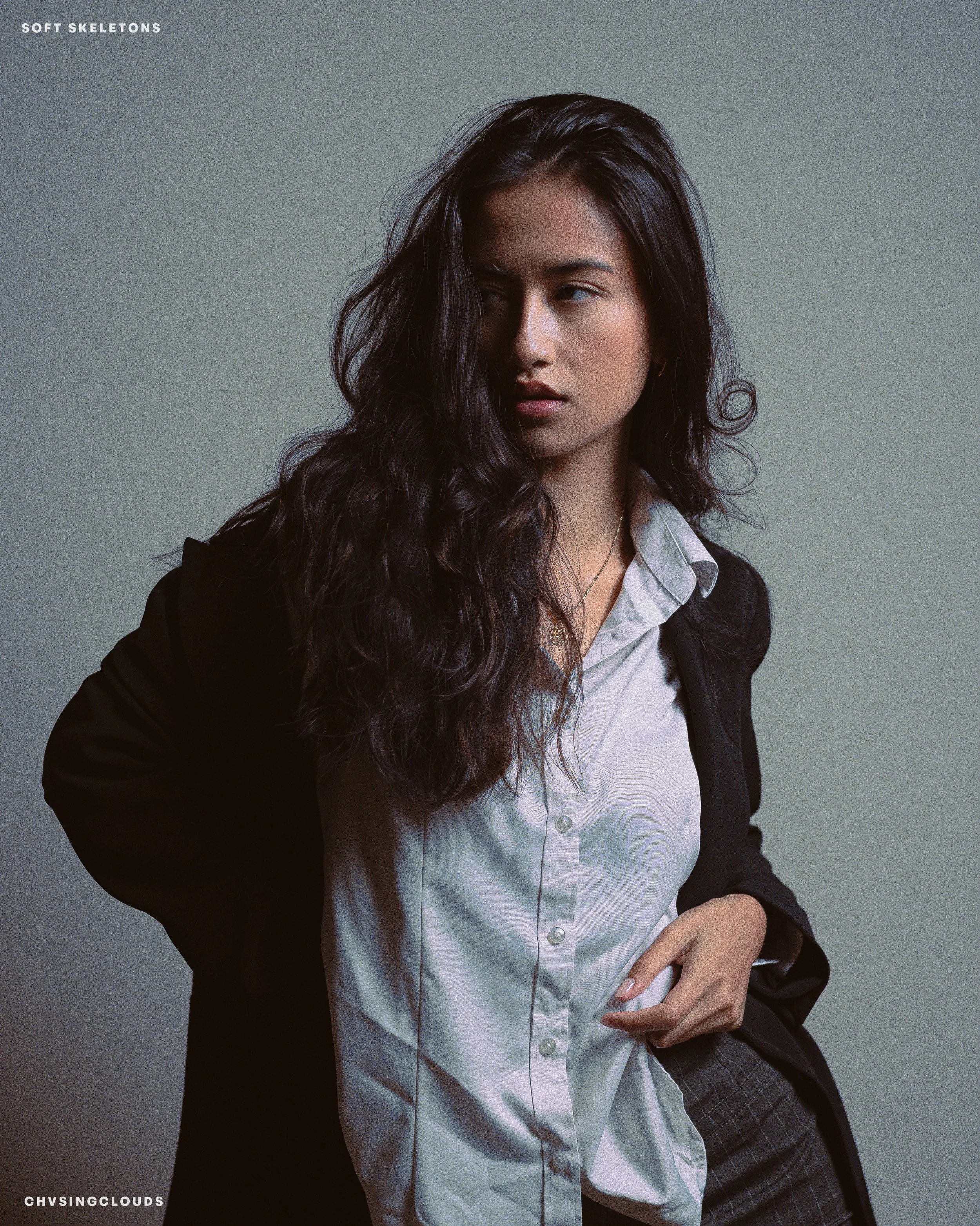 A woman with long, wavy dark hair, wearing a white button-up shirt and a black blazer, standing against a plain gray background.