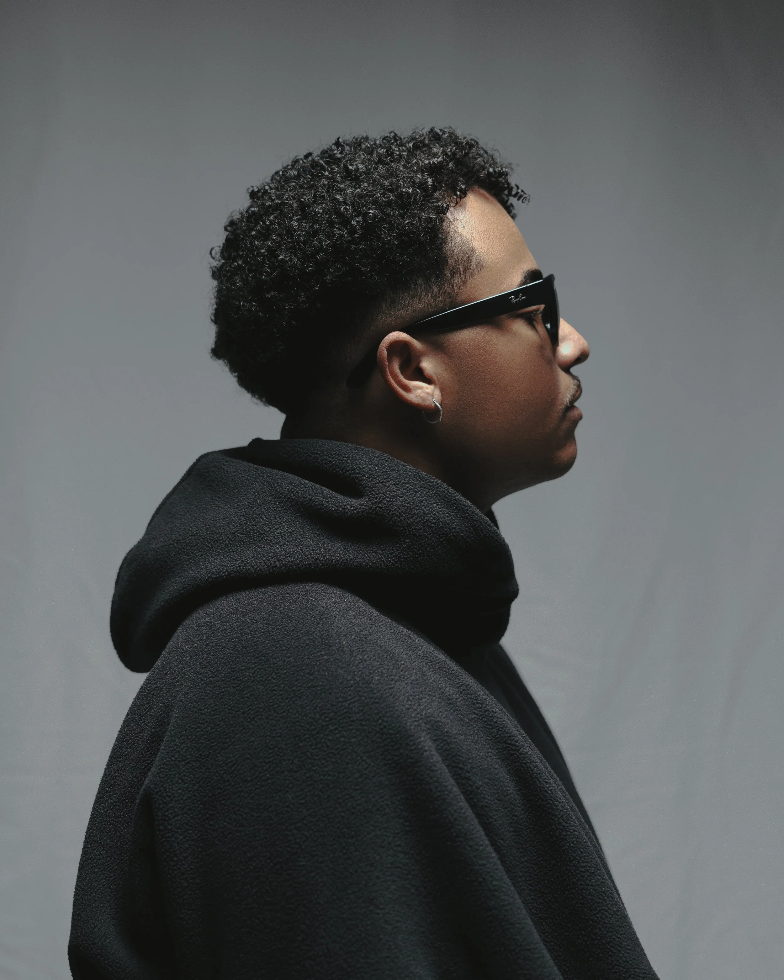 Side profile of a young person with curly hair, wearing black sunglasses and a black hoodie, against a plain gray background.