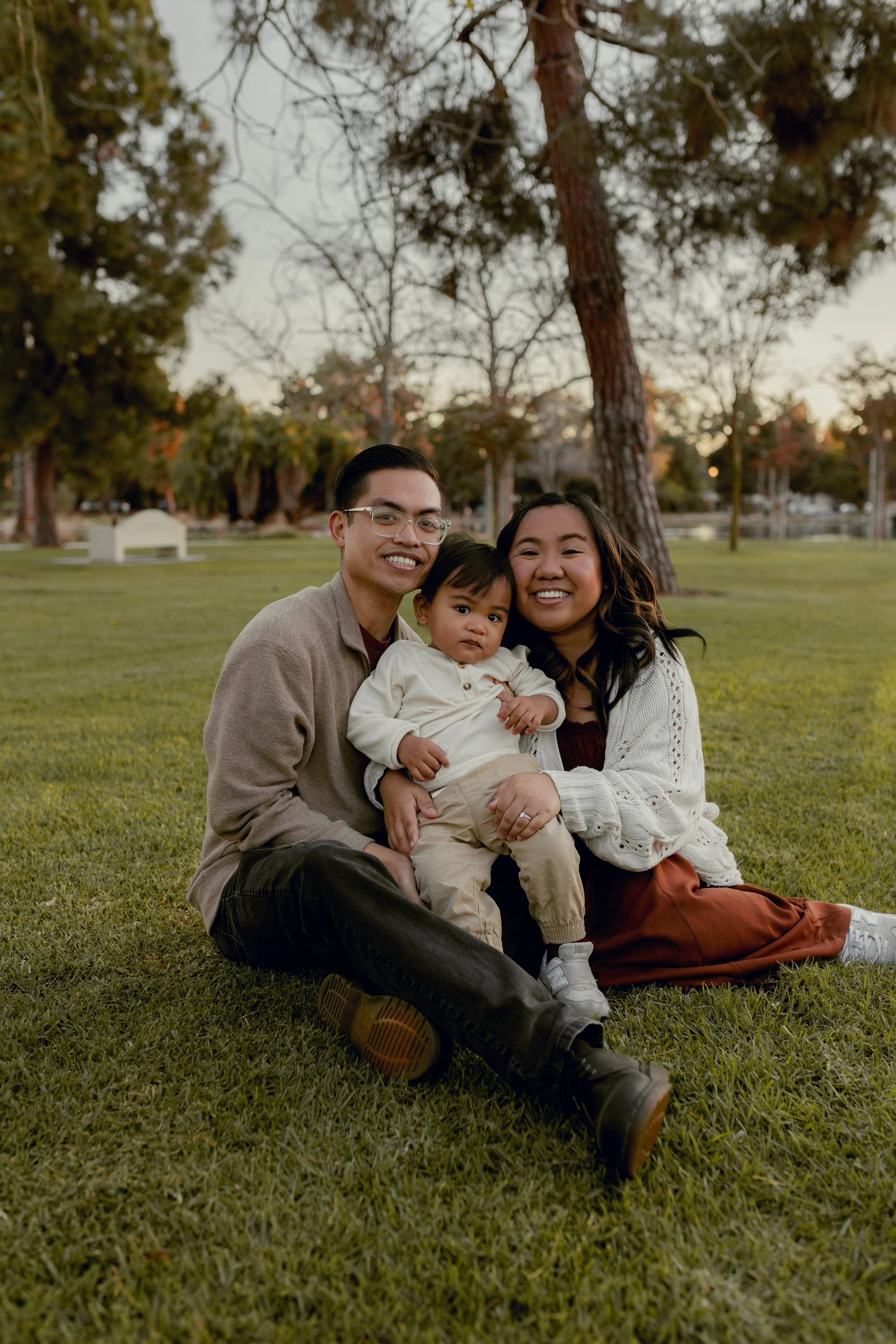 A happy family of three sitting on grass in a park during sunset, with trees and a few benches in the background.