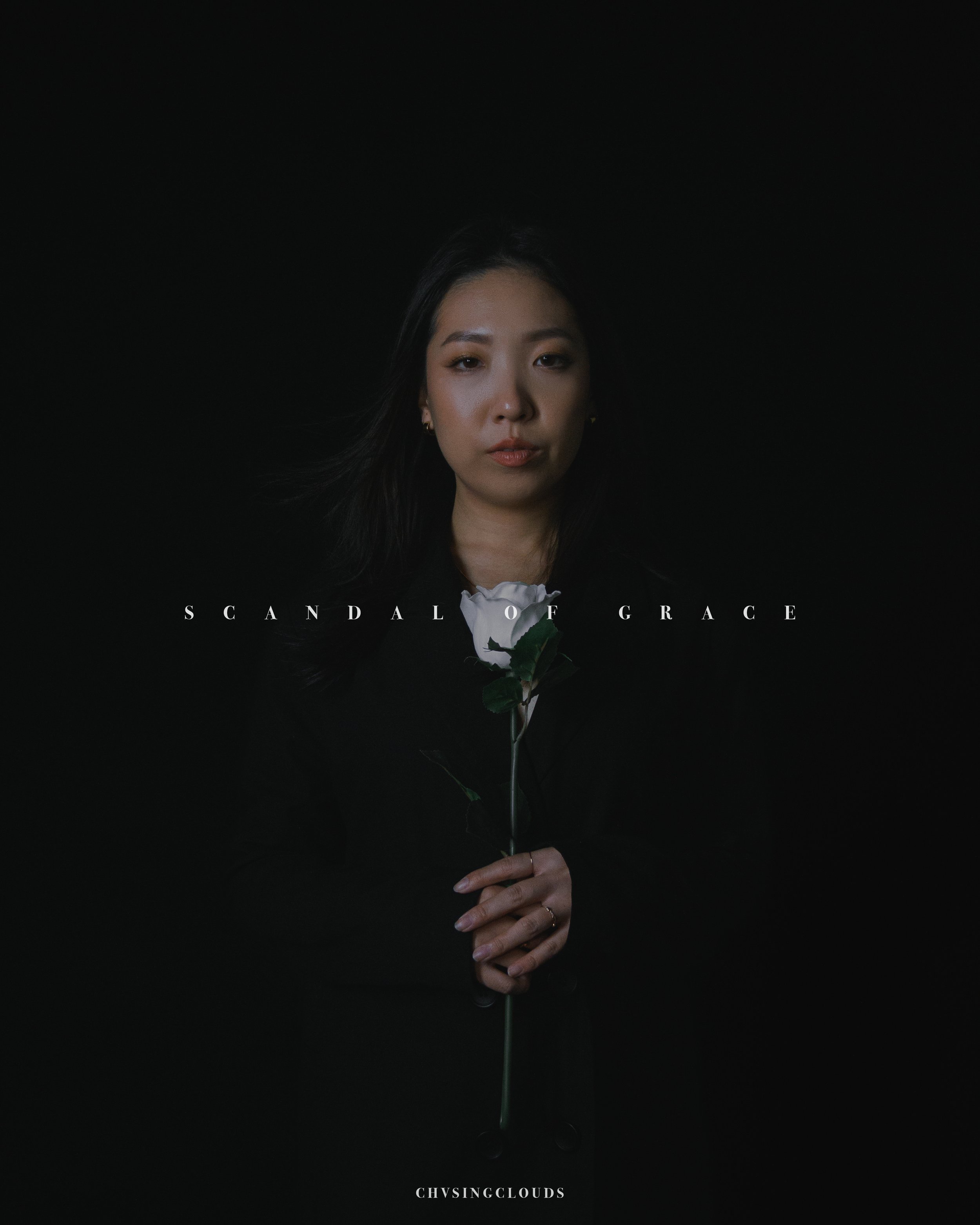 A woman with dark hair holding a white rose against a black background, with text reading 'Scandal of Grace' and 'Chvsingclouds'.