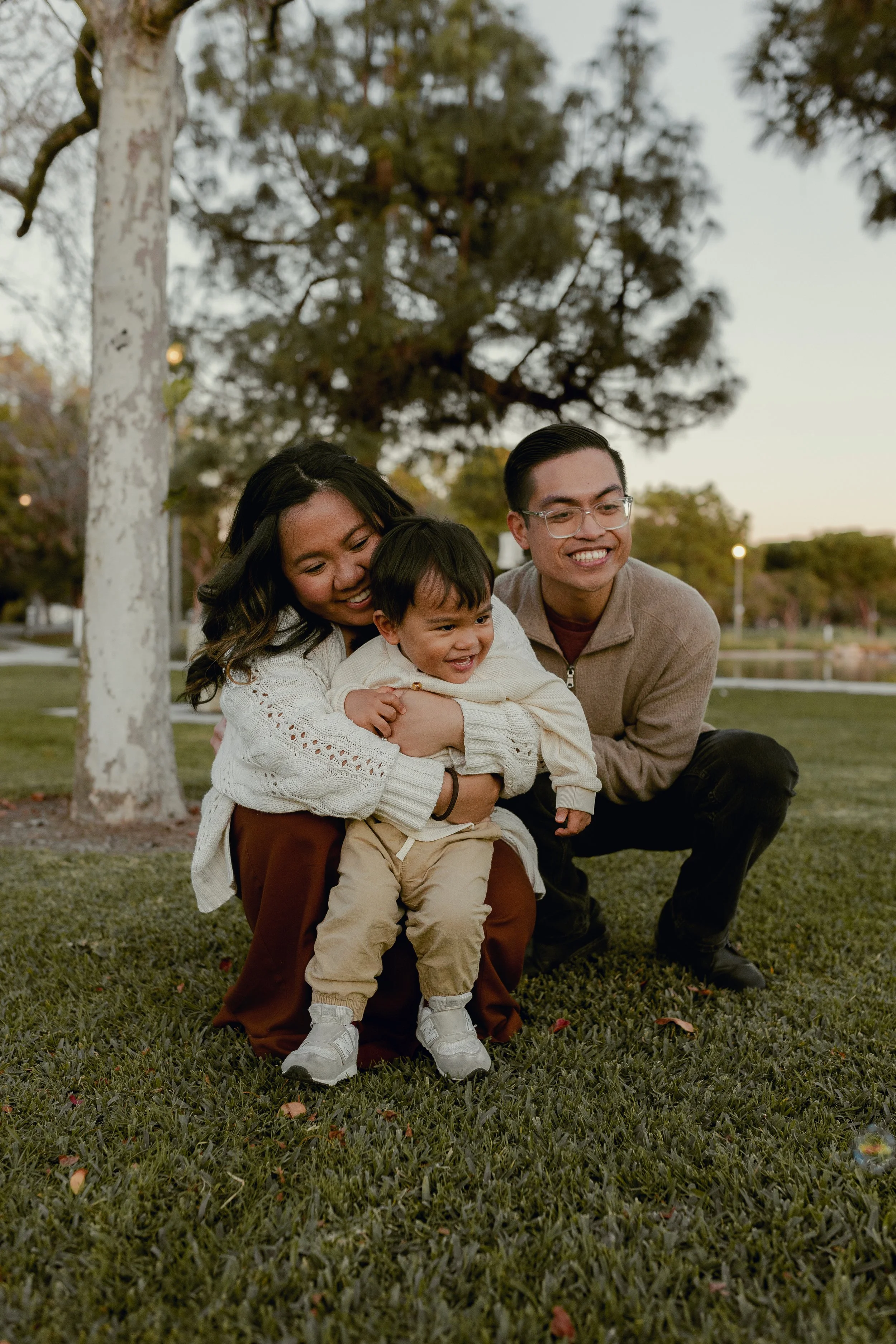 A family of three, including a woman, a young boy, and a man, are outdoors smiling and hugging each other on a grassy field with trees in the background during sunset.