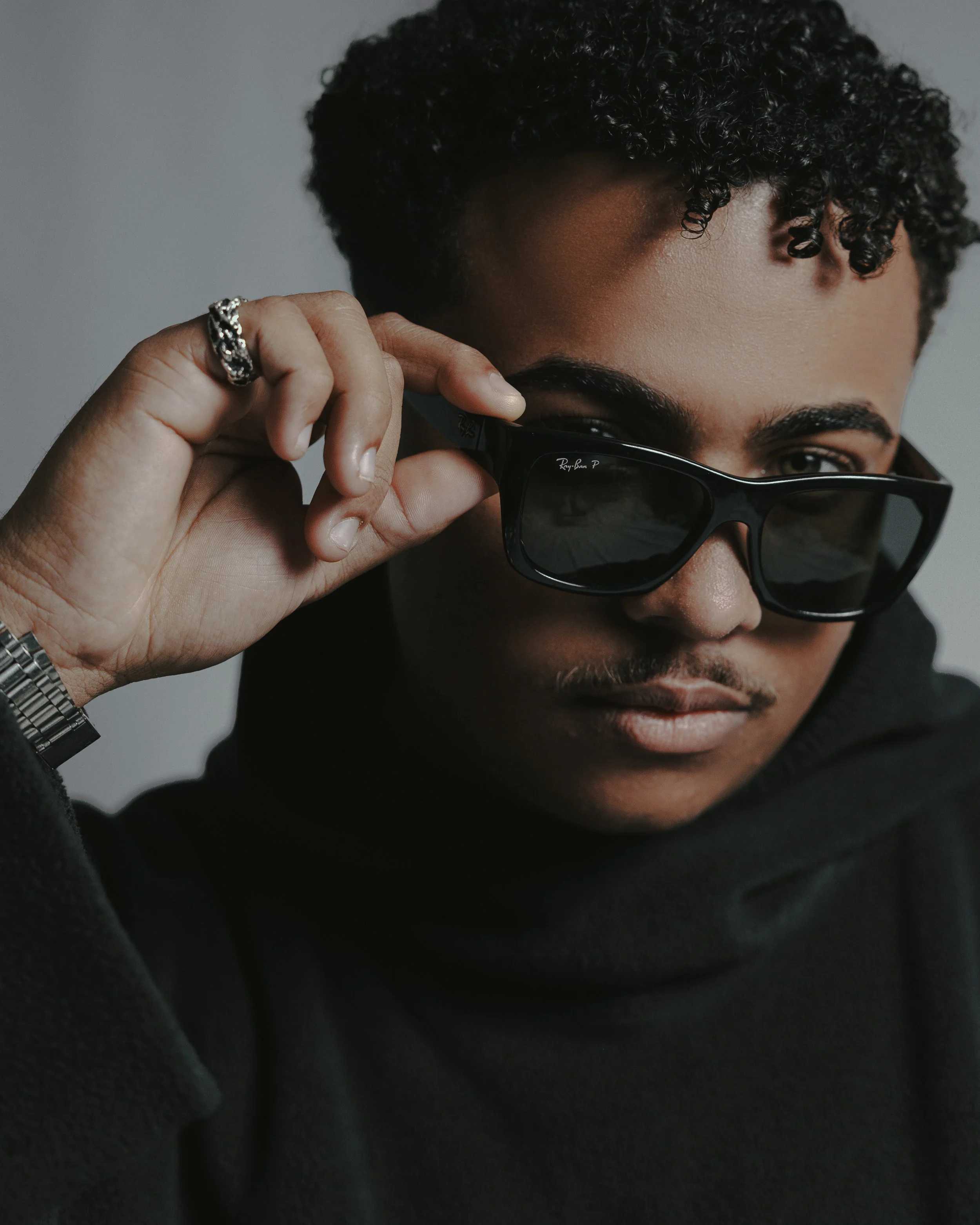 A person with curly hair wearing sunglasses and a black hoodie, holding the sunglasses with their left hand, which has rings and a watch.