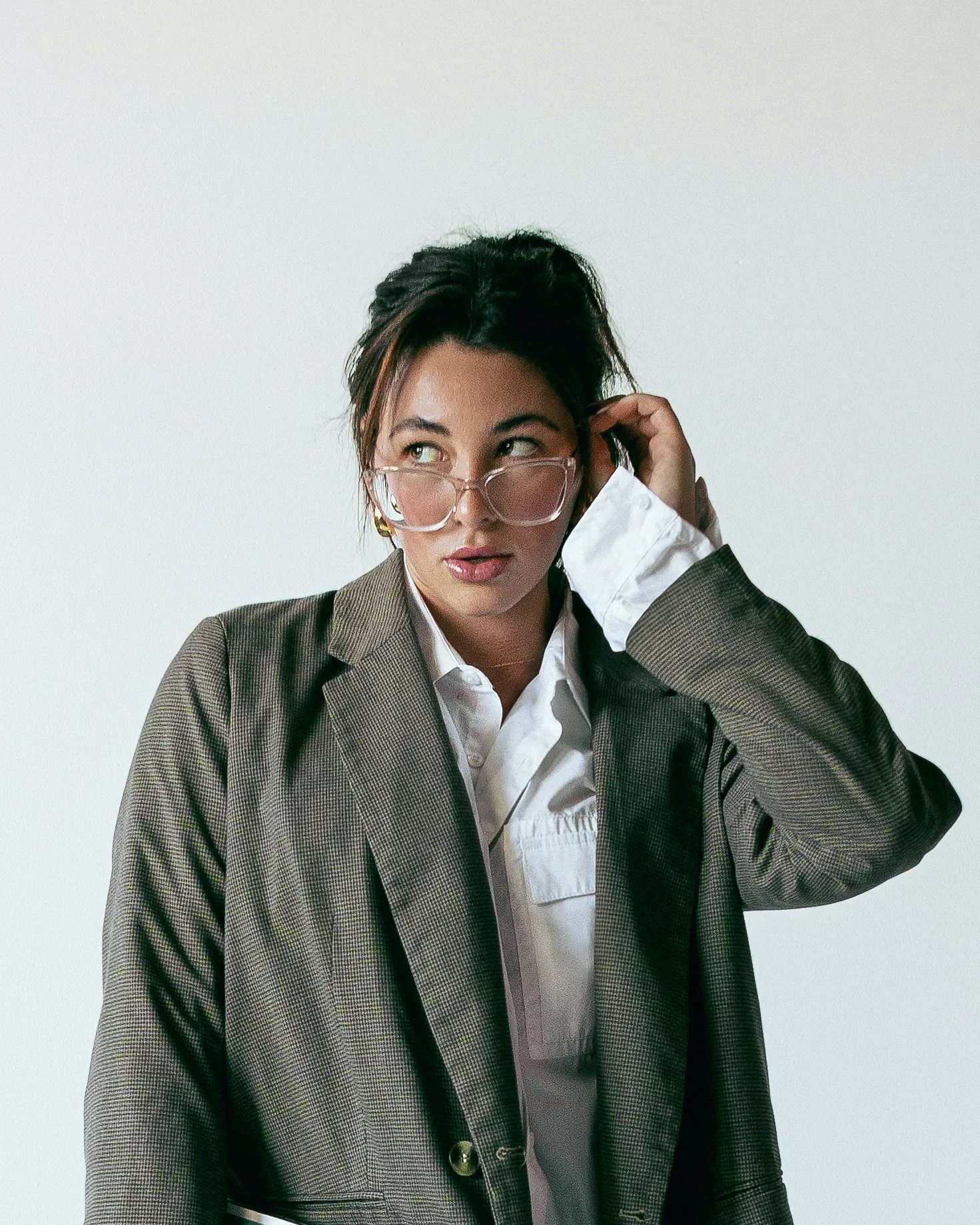 A woman with dark hair, glasses, and earrings, wearing a white shirt and a brown blazer, looks to the side while adjusting her glasses against a plain white background.
