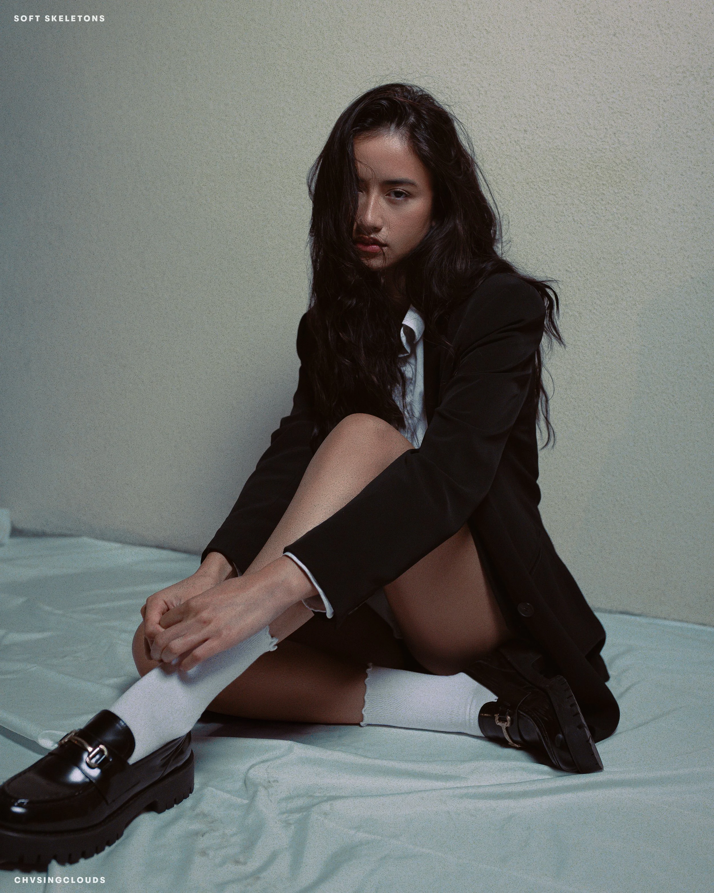 Woman with long dark hair sitting on a white sheet, wearing a black blazer, white shirt, white socks, and black loafers. She is against a plain beige wall, gazing at the camera with a serious expression.