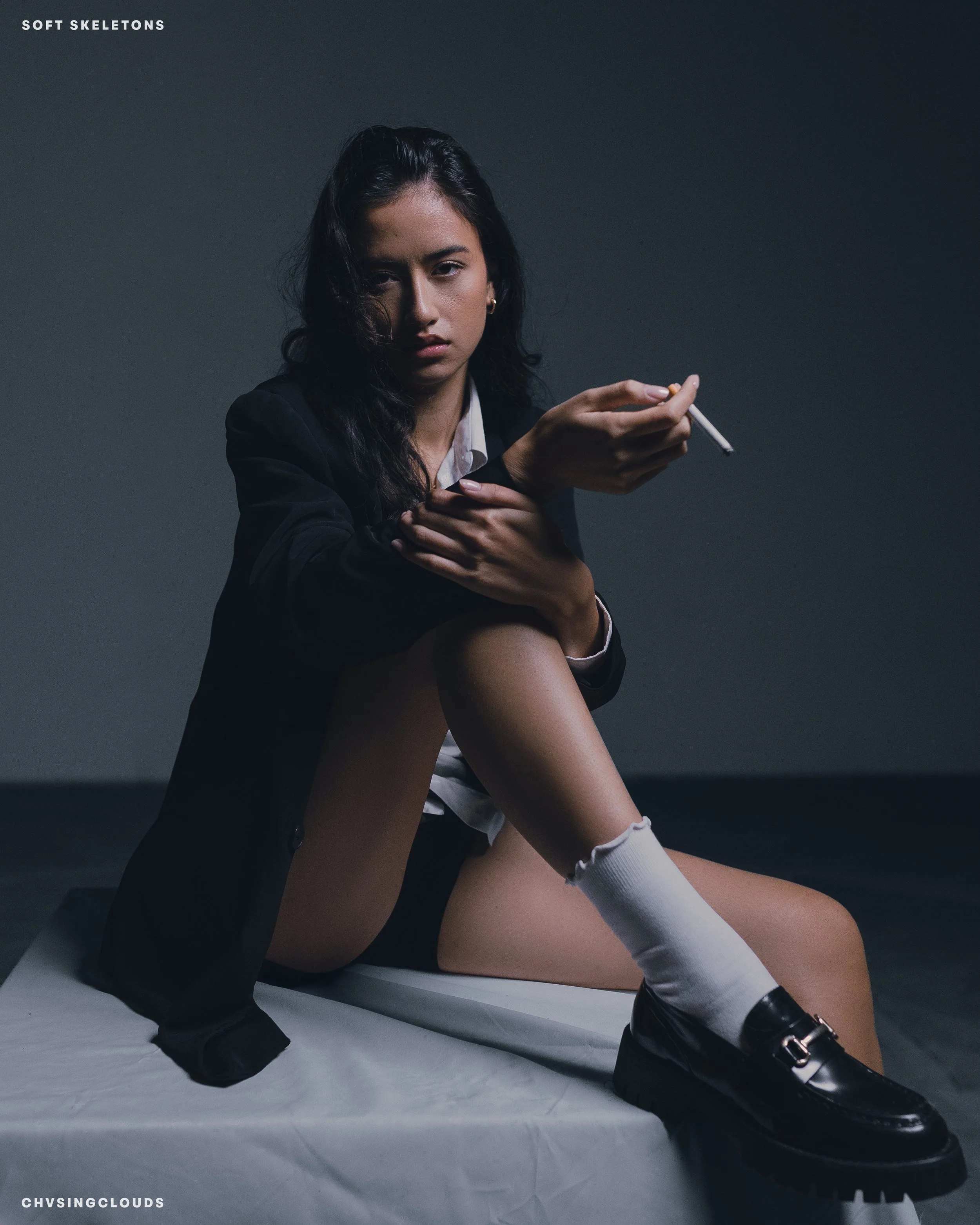 A young woman with long dark hair sitting on a white sheet, wearing a black blazer, white shirt, black shoes, and knee-high white socks, holding a cigarette in her right hand, with a serious expression against a plain dark background.