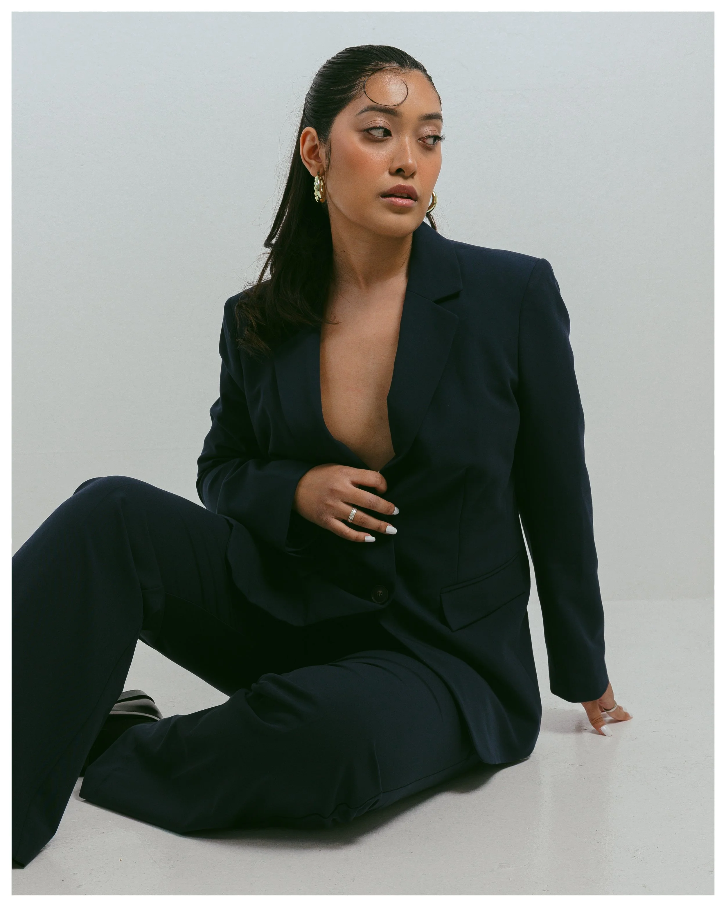 A woman sitting on the floor against a plain light gray wall, dressed in a dark blazer and pants, with her hair slicked back, wearing large earrings, and looking to her left with a serious expression.