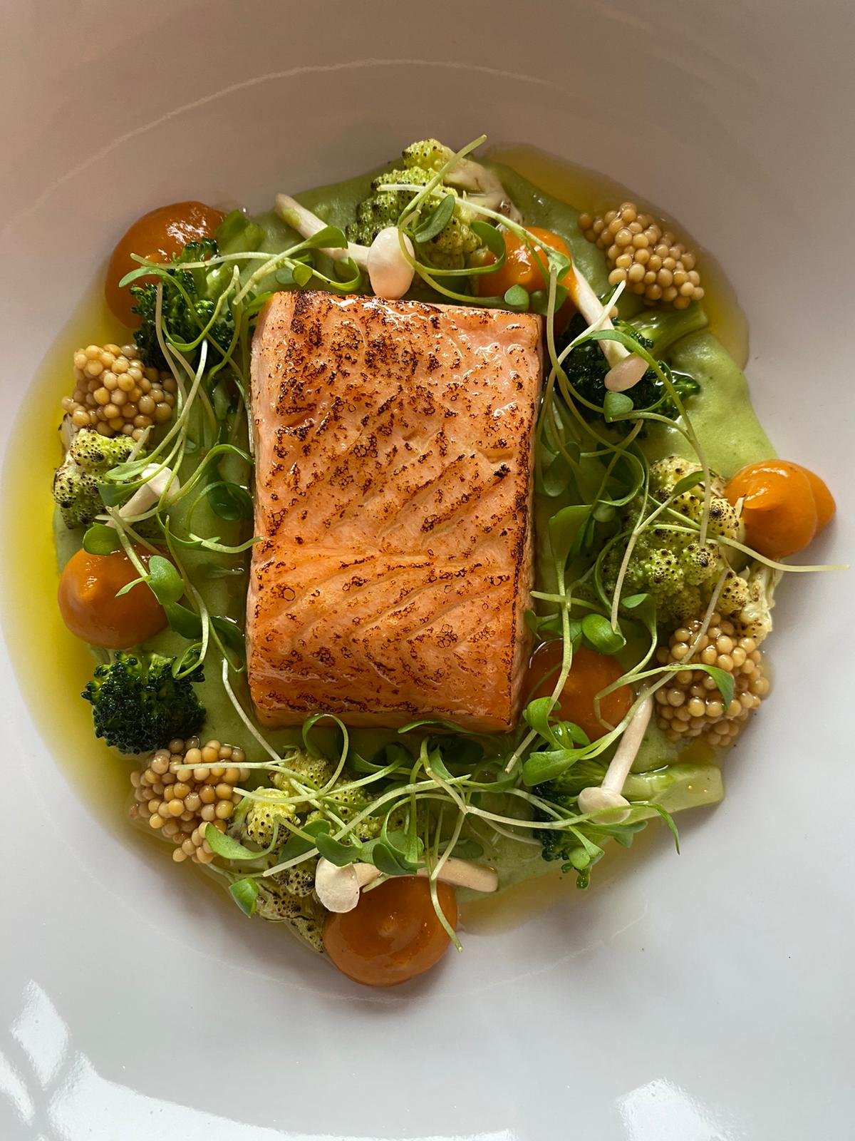 Pan-seared salmon fillet on a bed of green vegetables and microgreens, garnished with cherry tomatoes and mustard seeds.