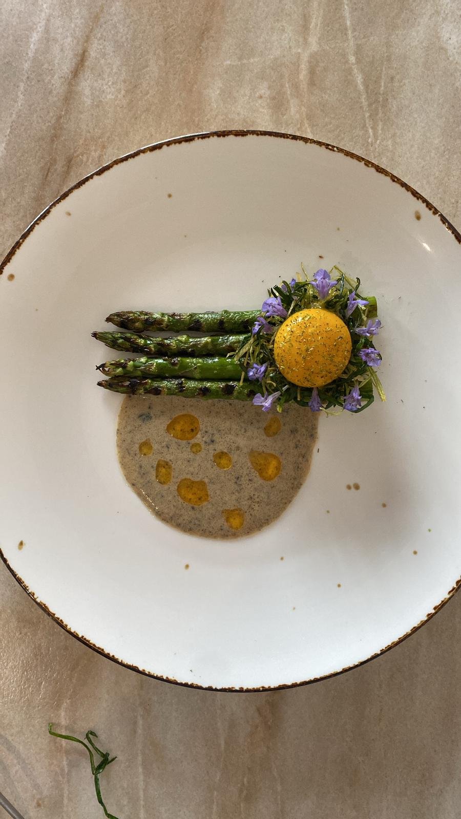 A plate with roasted green asparagus spears, topped with edible purple flowers and a yellow, round garnish. There is a light-colored sauce with small drops of oil or sauce around it on the plate.