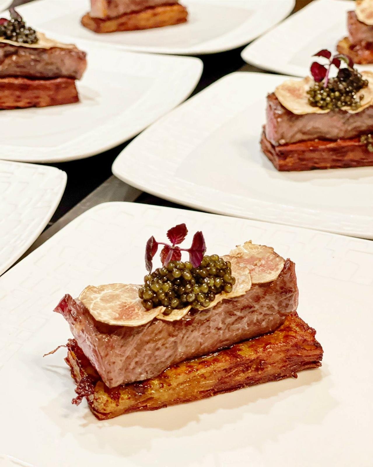 A plated dish featuring a thick slice of cooked meat on a bed of crispy potato or pastry, topped with black caviar, thin slices of garlic, small purple leaves, and garnished with a tiny sprig of fresh herbs.