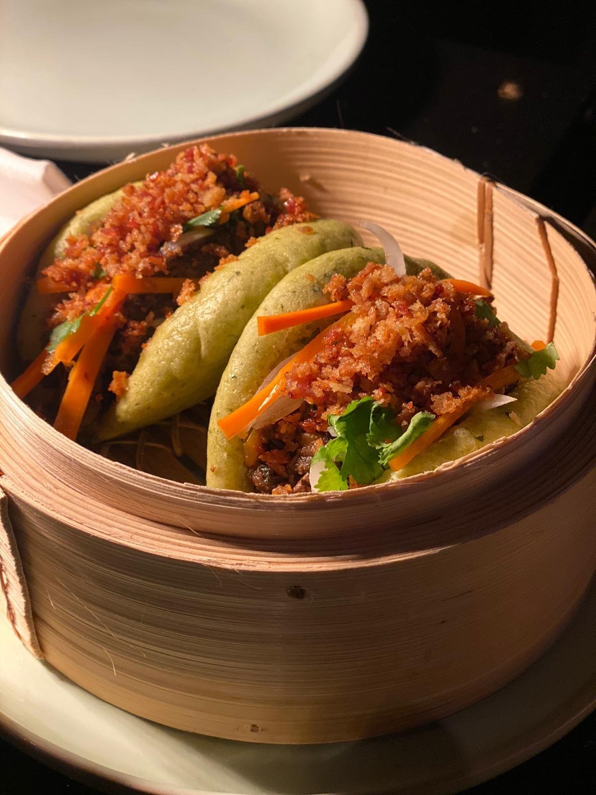 Three steamed bao buns filled with minced meat, shredded carrots, and cilantro, served in a round bamboo steamer basket.