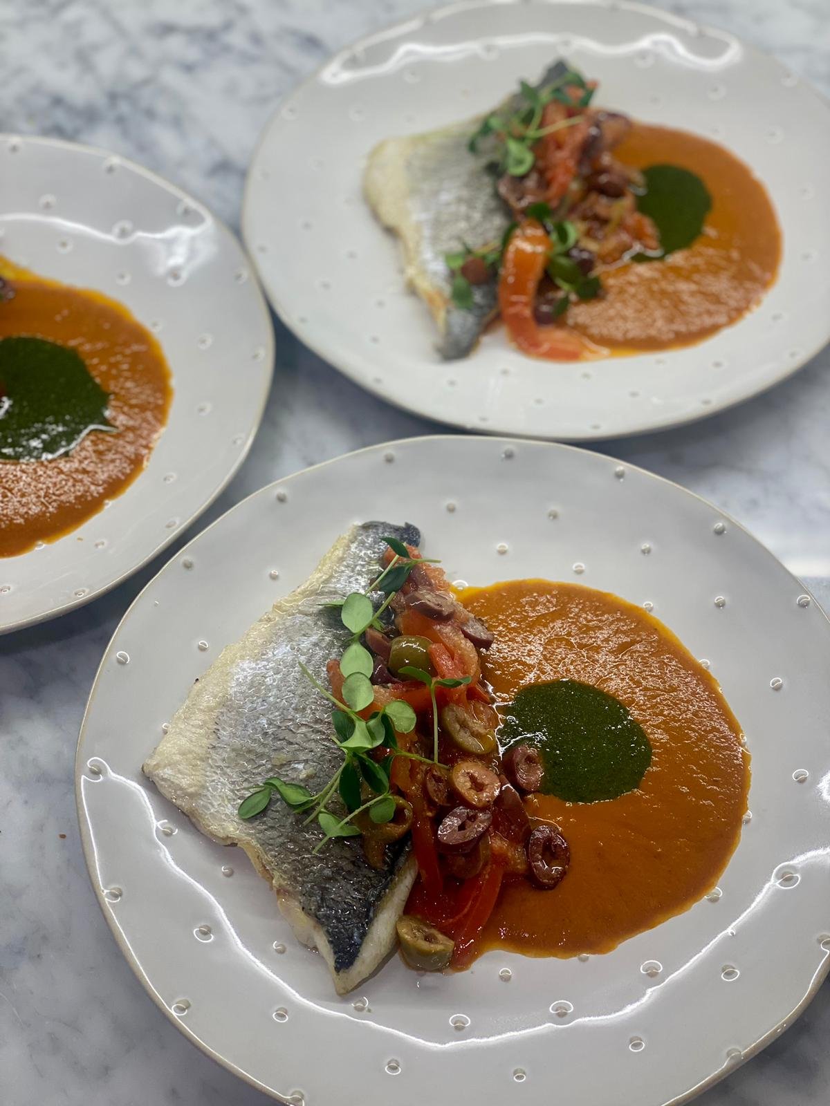 Three white plates with silver fish fillets, each topped with sautéed vegetables and garnished with microgreens, served with a side of orange sauce and green sauce garnished with a small green herb.