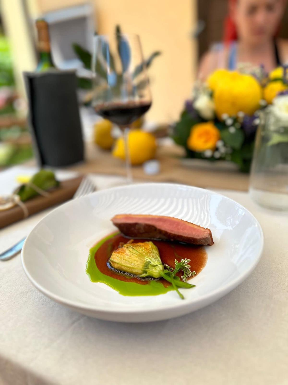 Close-up of a fine dining plate with a slice of cooked meat, a roasted vegetable, and colorful sauces, with a blurred background showing a wine glass, flowers, and a person.