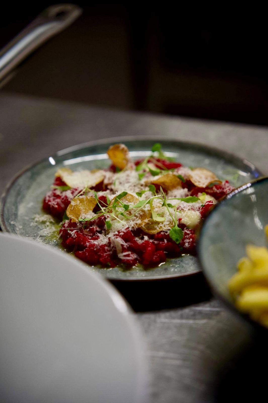 Close-up of a plated dish with red meat tartare garnished with microgreens, grated cheese, and crispy chips, served on a dark plate.