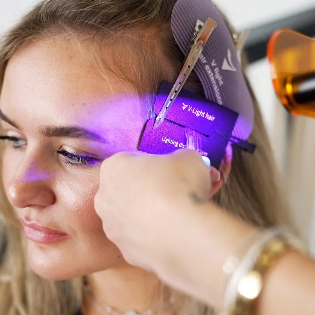 A woman undergoing a hair laser treatment, with a technician using a device emitting purple light on her forehead.