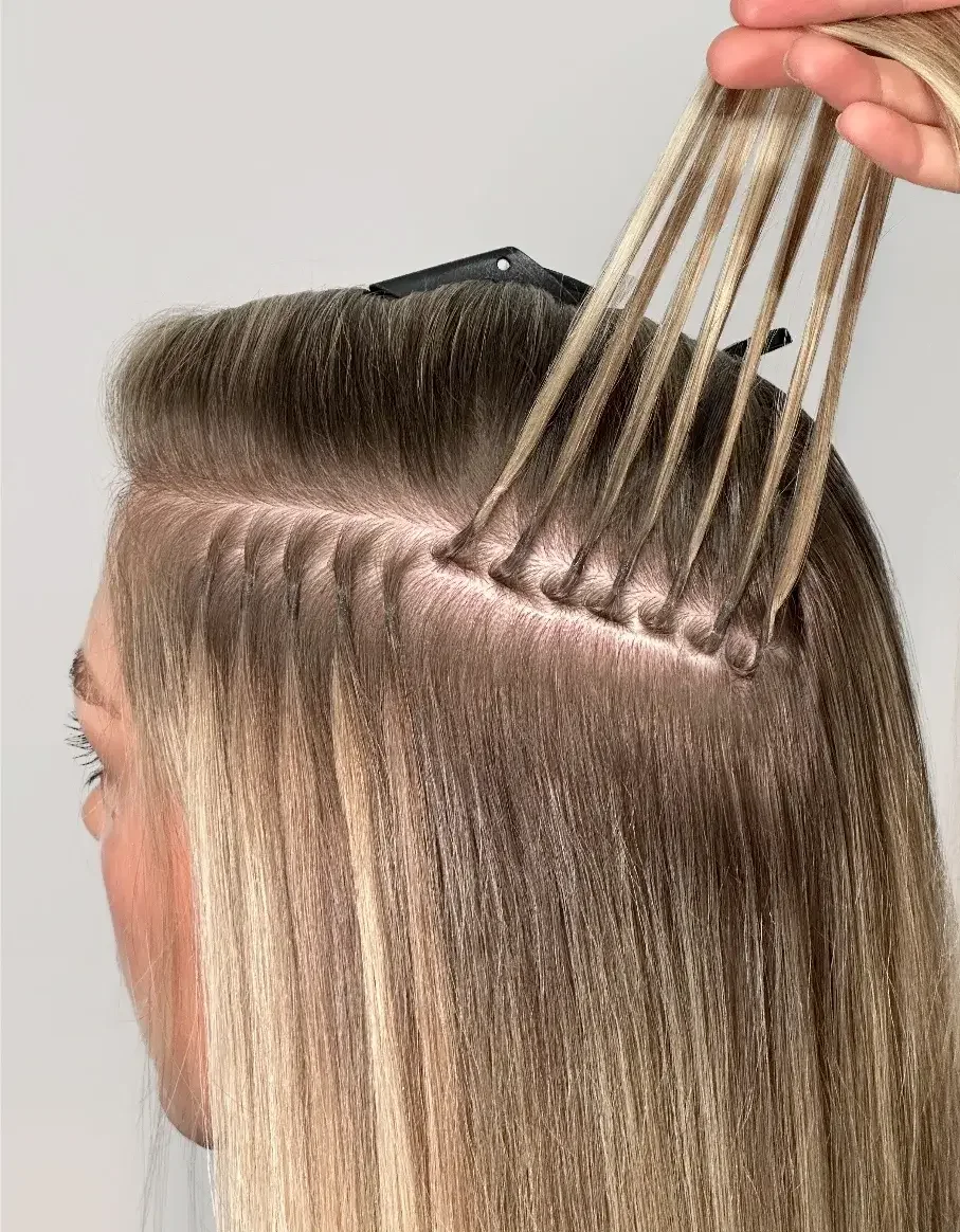 A hairstylist applying hair dye or bleach to a woman's long, straight blonde hair, sectioned and held with clips.
