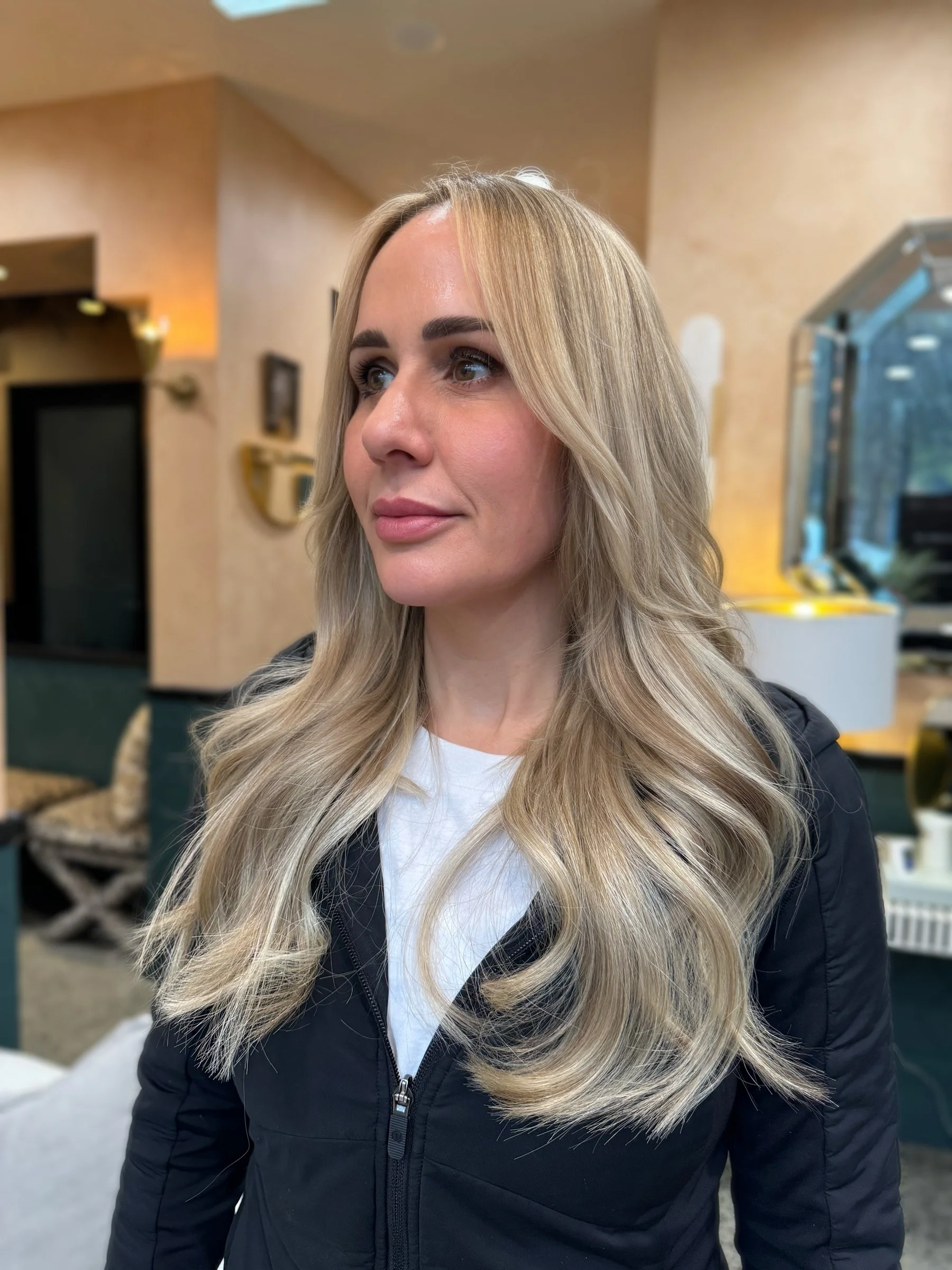 Close-up of a woman with long, wavy blonde hair, wearing a black jacket and white shirt, standing indoors with a blurred background.