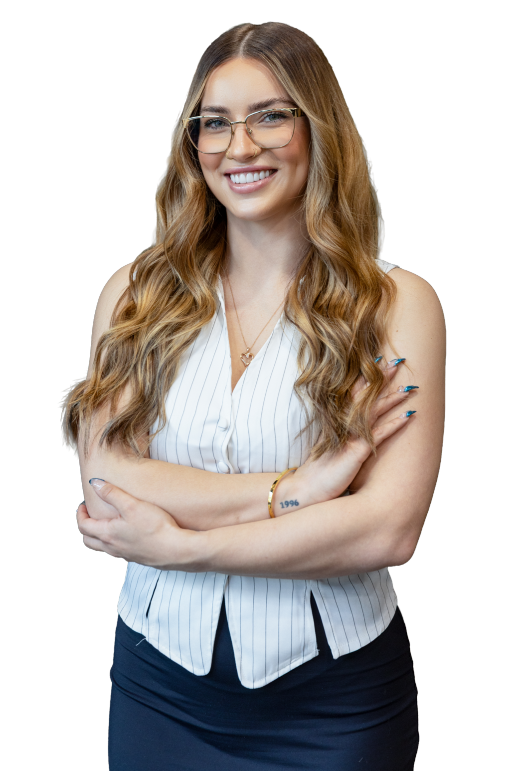 A smiling woman with long wavy red hair, wearing glasses, a white sleeveless blouse with vertical stripes, and a gold bracelet. She is standing with arms crossed and has a small tattoo on her left wrist that reads '1996'.