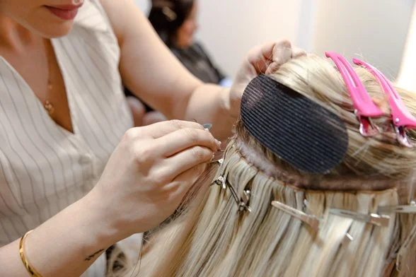 A hair stylist is applying a hair treatment or extension to a client's blonde hair, which is clipped in rollers and held with metal clips.