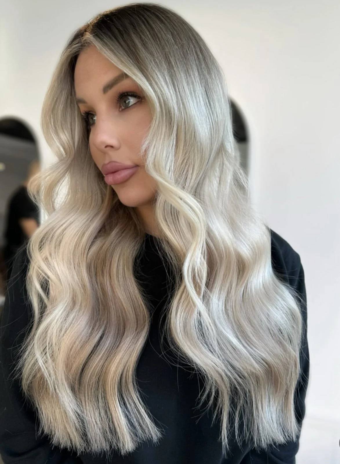 A woman with long, wavy blonde hair with dark roots, wearing a black top, looking to her left in a salon setting.