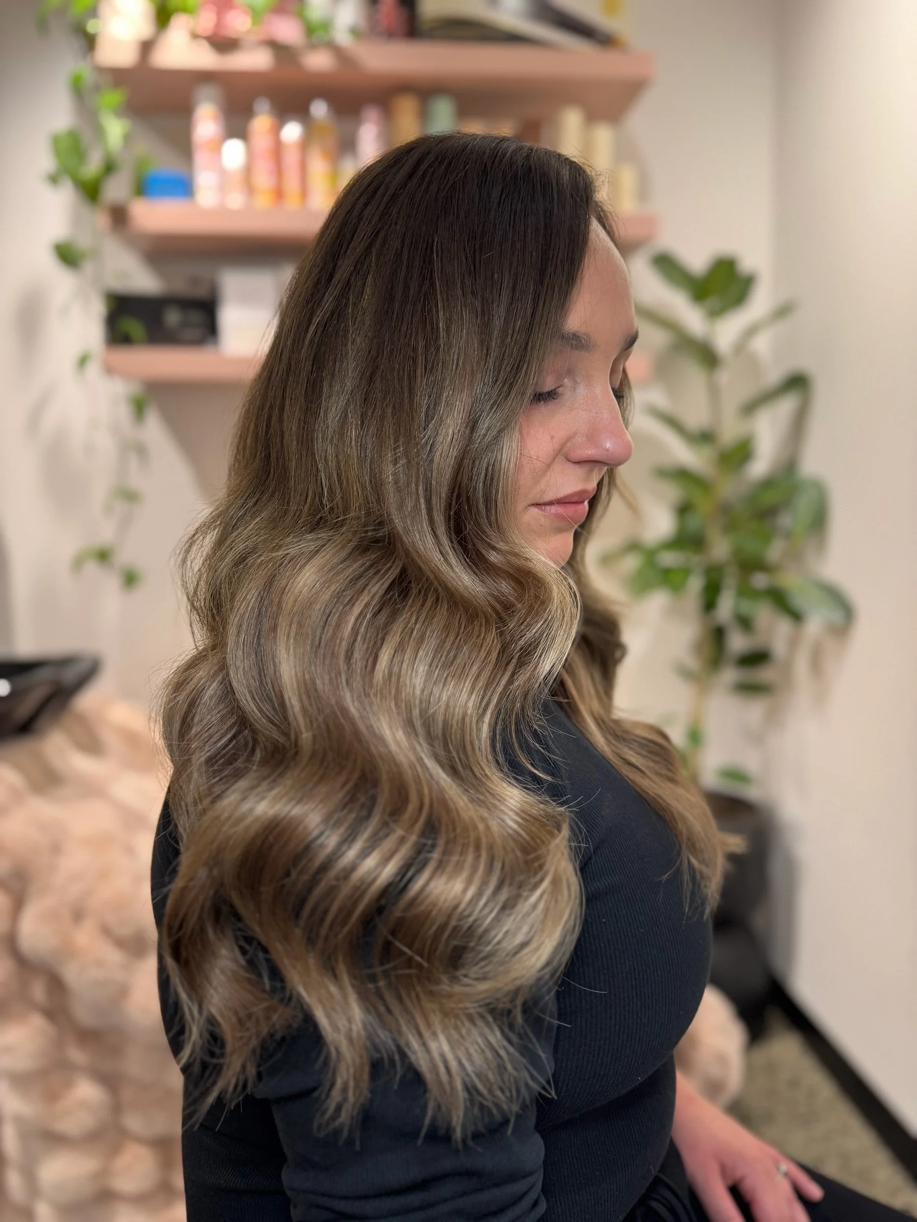 A woman with long wavy hair in shades of brown and blonde, with highlights, sitting with her eyes closed inside a room with shelves and plants in the background.