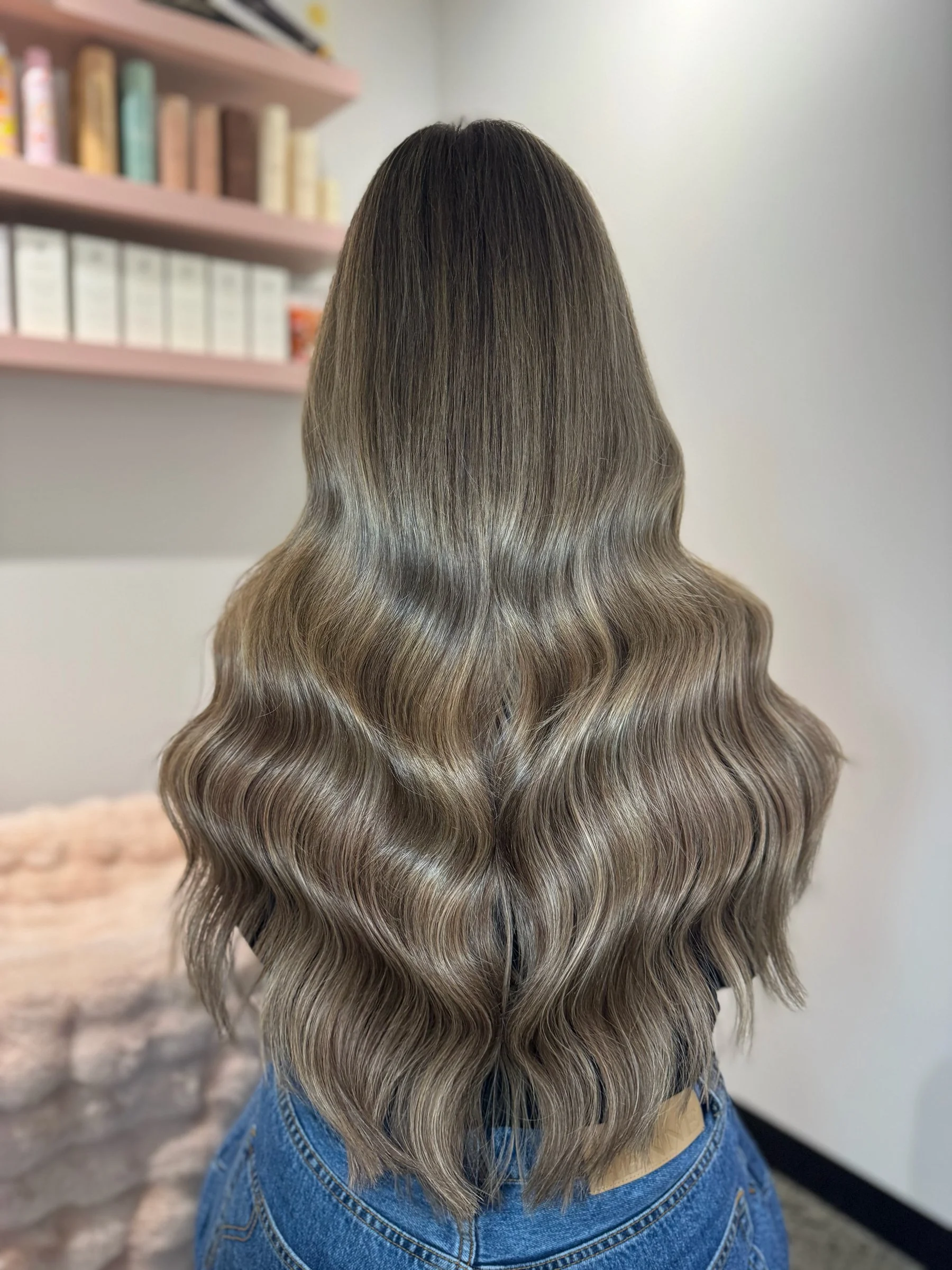 Back view of a woman with long, wavy hair that is dark at the roots and transitions to ash blonde tips, wearing a blue denim jacket.