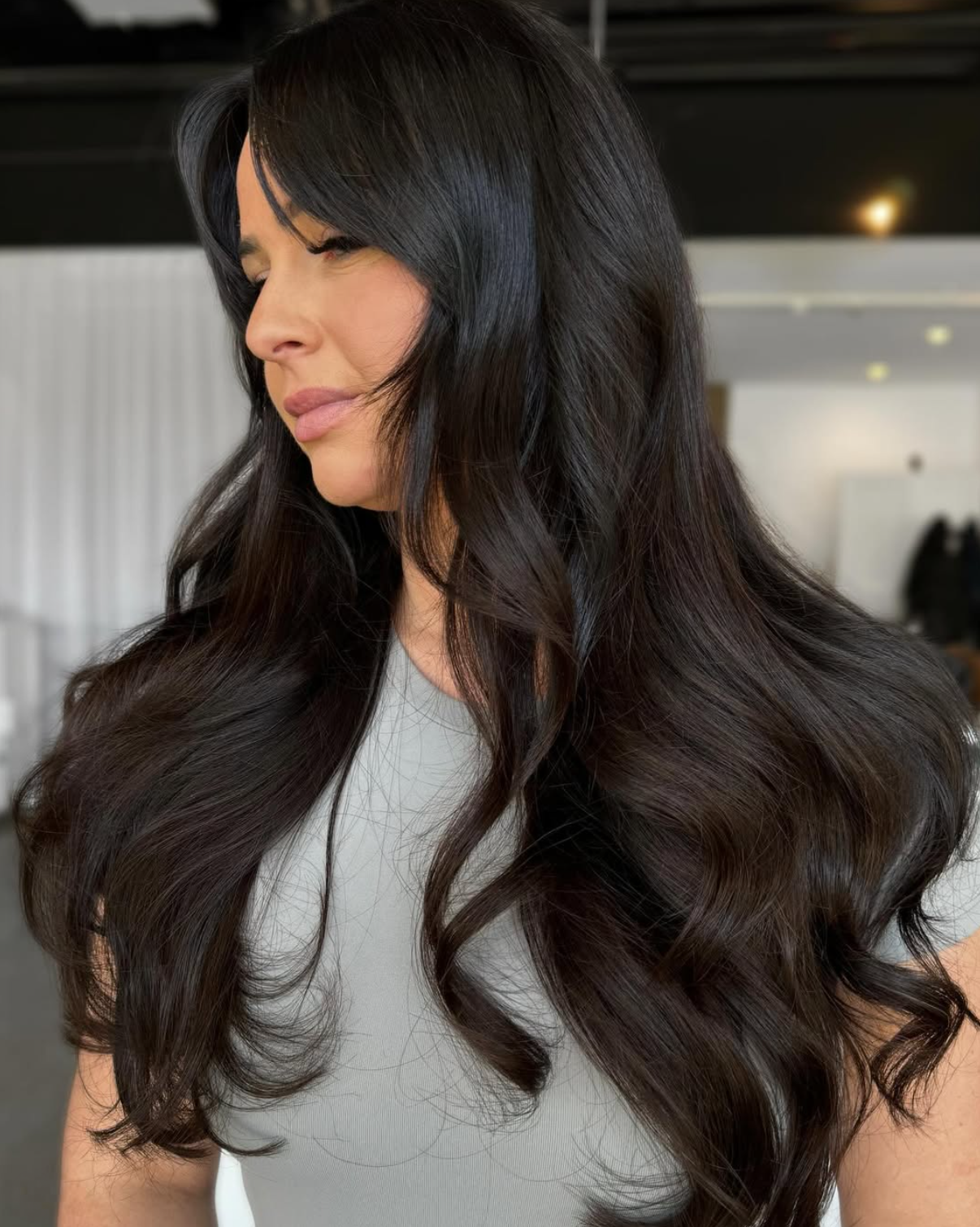 A woman with long, dark, wavy hair looking to the side in a well-lit indoor setting.