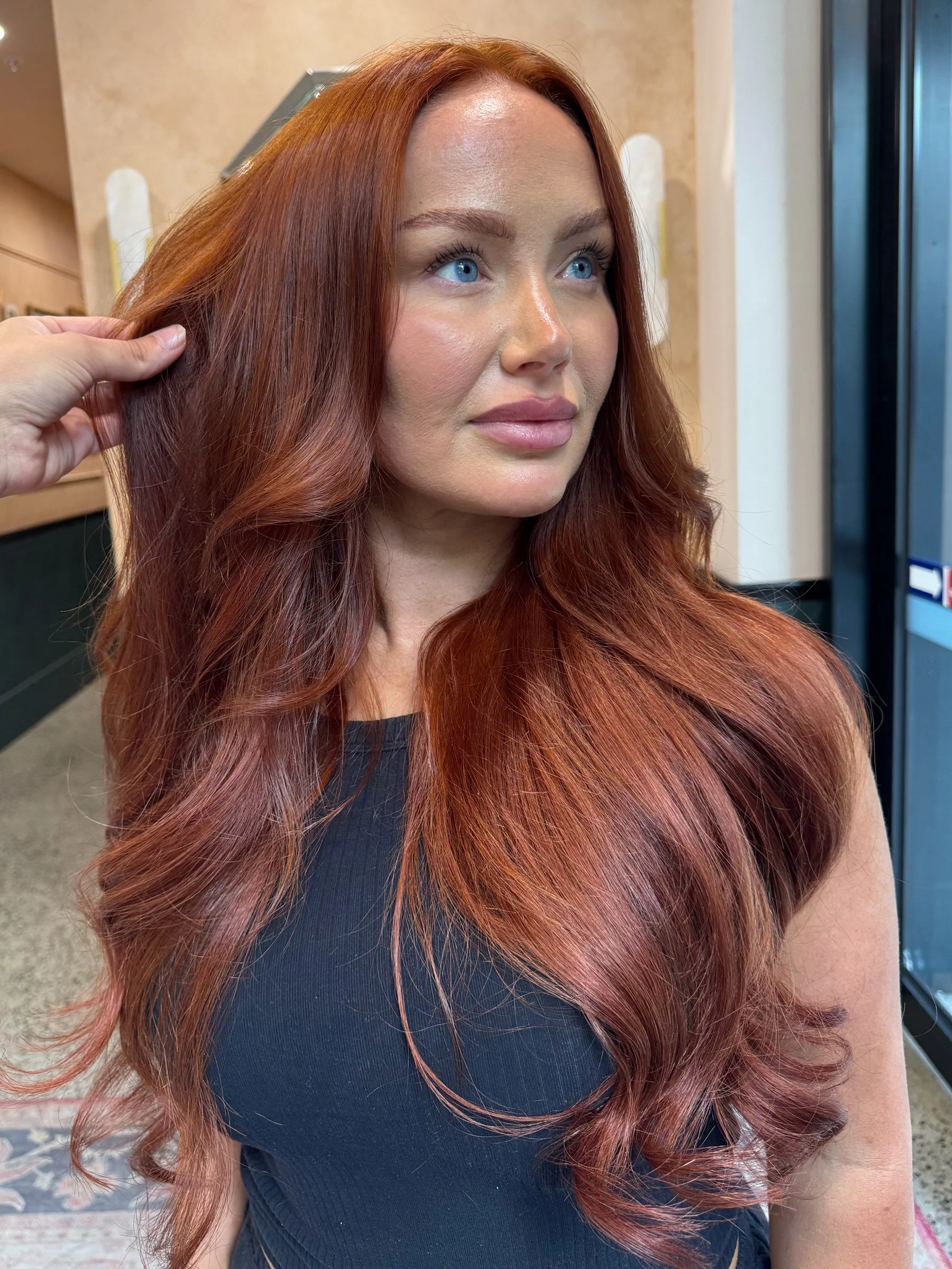 Close-up of a woman with long, wavy red hair and blue eyes, wearing a black top, in a salon or similar setting.