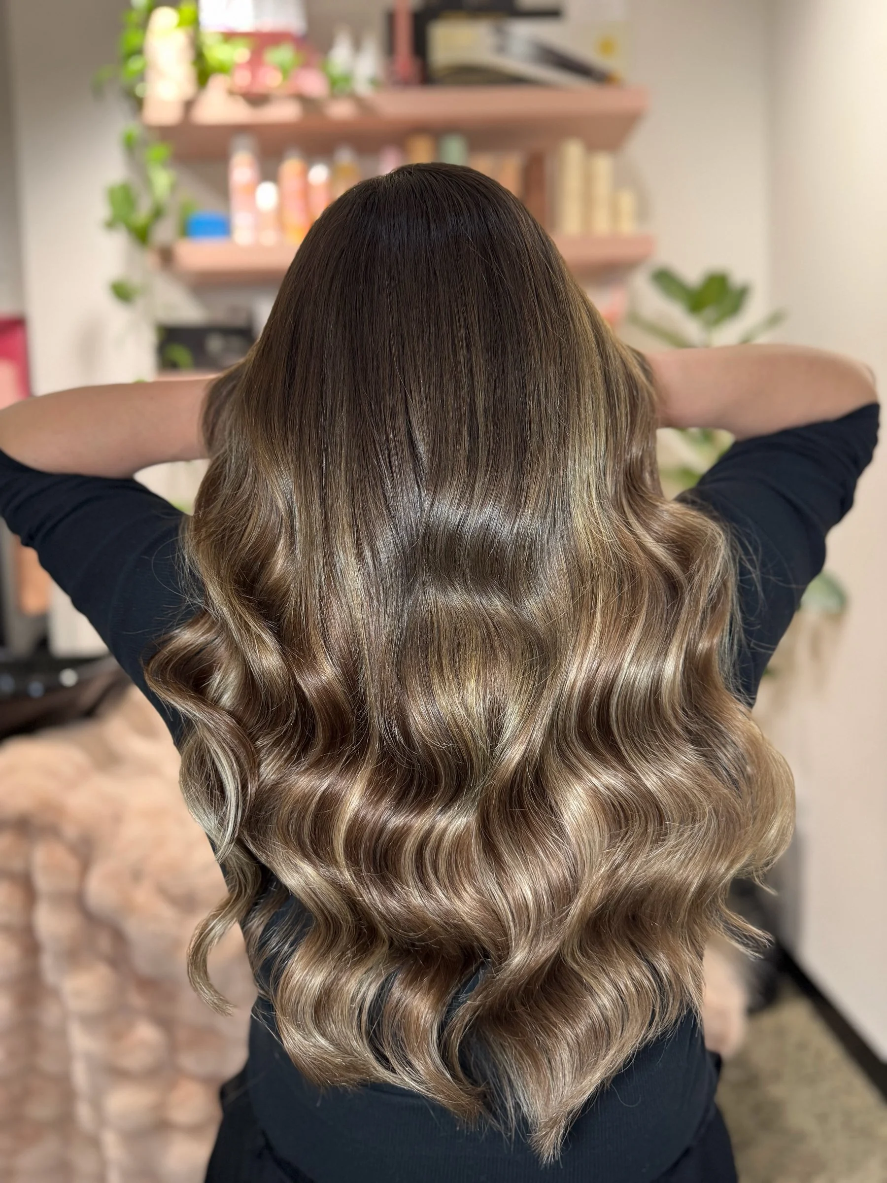 Woman with long, wavy, ombre hair, transition from dark brown roots to lighter blonde tips, sitting indoors with her back to the camera, arms resting behind her head, background includes shelves with beauty products and plants.