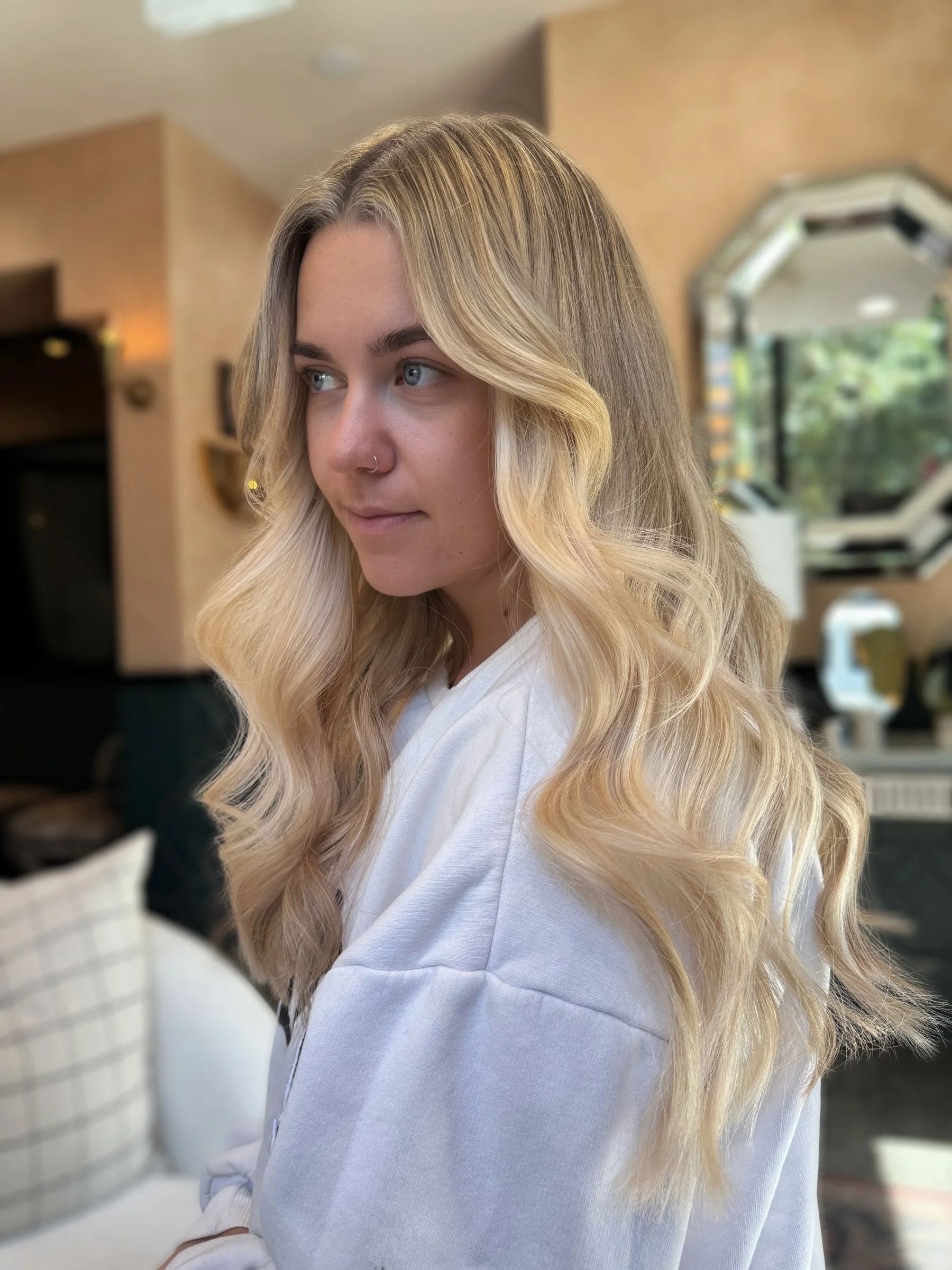 A young woman with long, wavy blonde hair and blue eyes, wearing a white top, is looking slightly to the left in a brightly-lit room with beige walls, a mirror, and blurred furniture in the background.