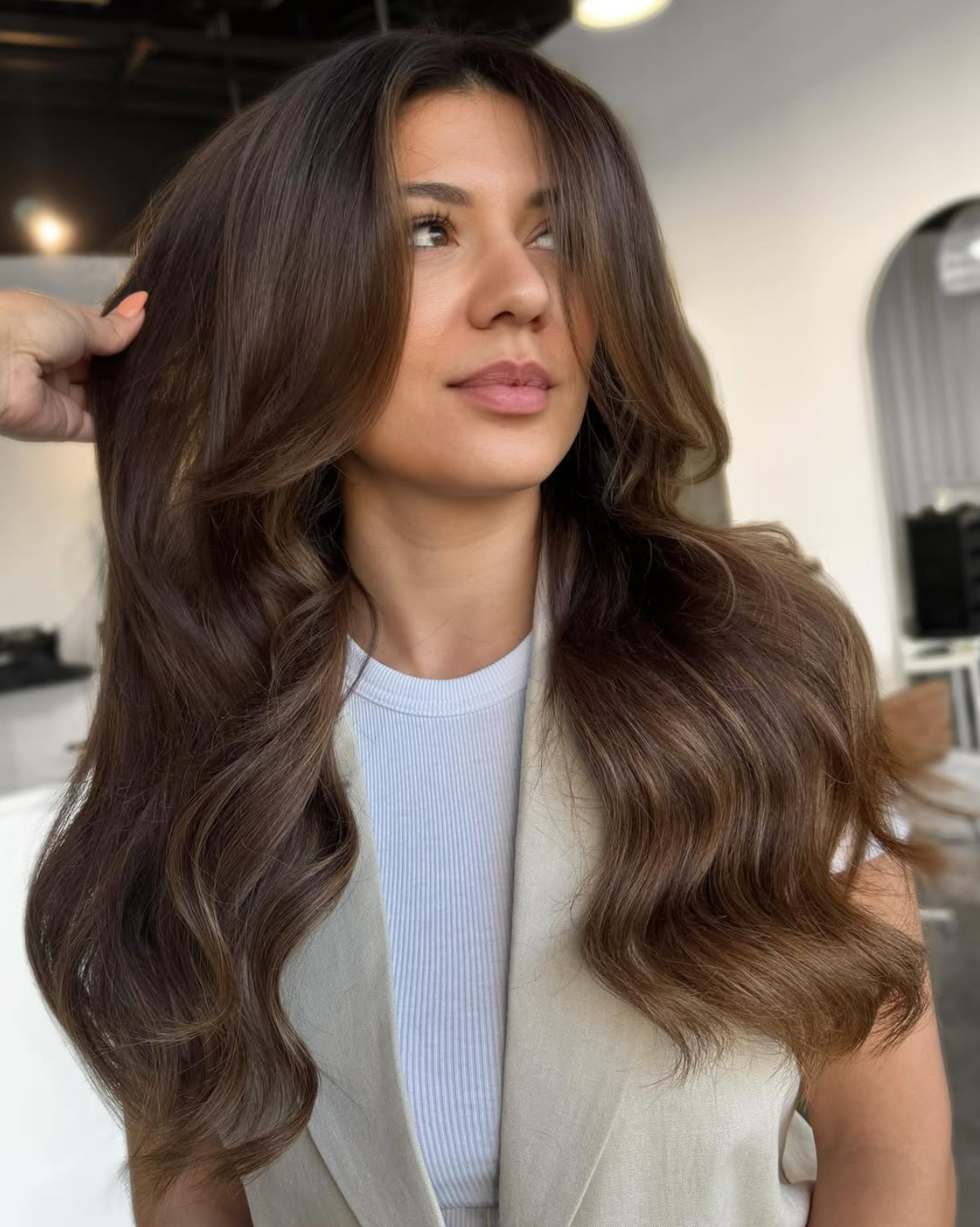 A woman with long, wavy brown hair looking to the side at a hairdresser, wearing a white top and a beige vest in a hair salon.