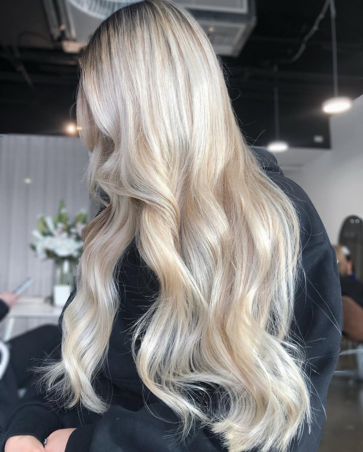 Blonde woman with long, wavy hair sitting indoors, with a blurred background showing flowers and a person using a phone.