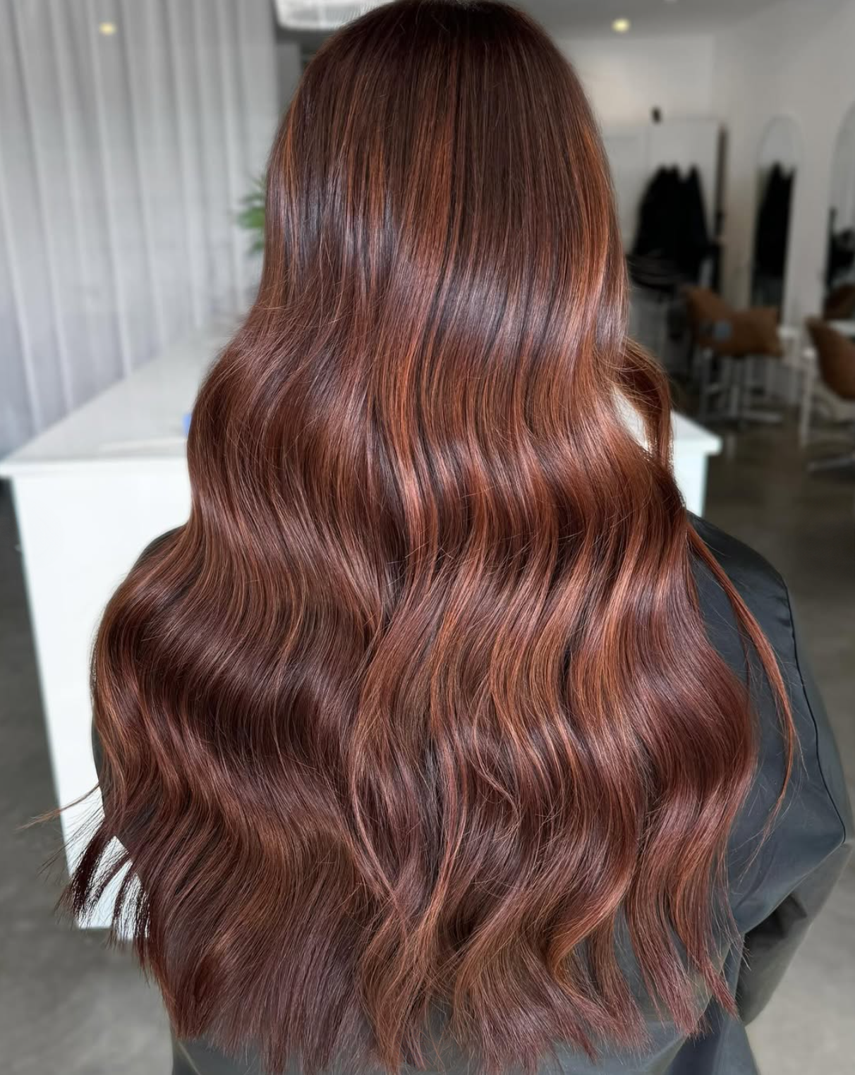 Back of a person with long, voluminous, wavy reddish-brown hair styled with loose waves.