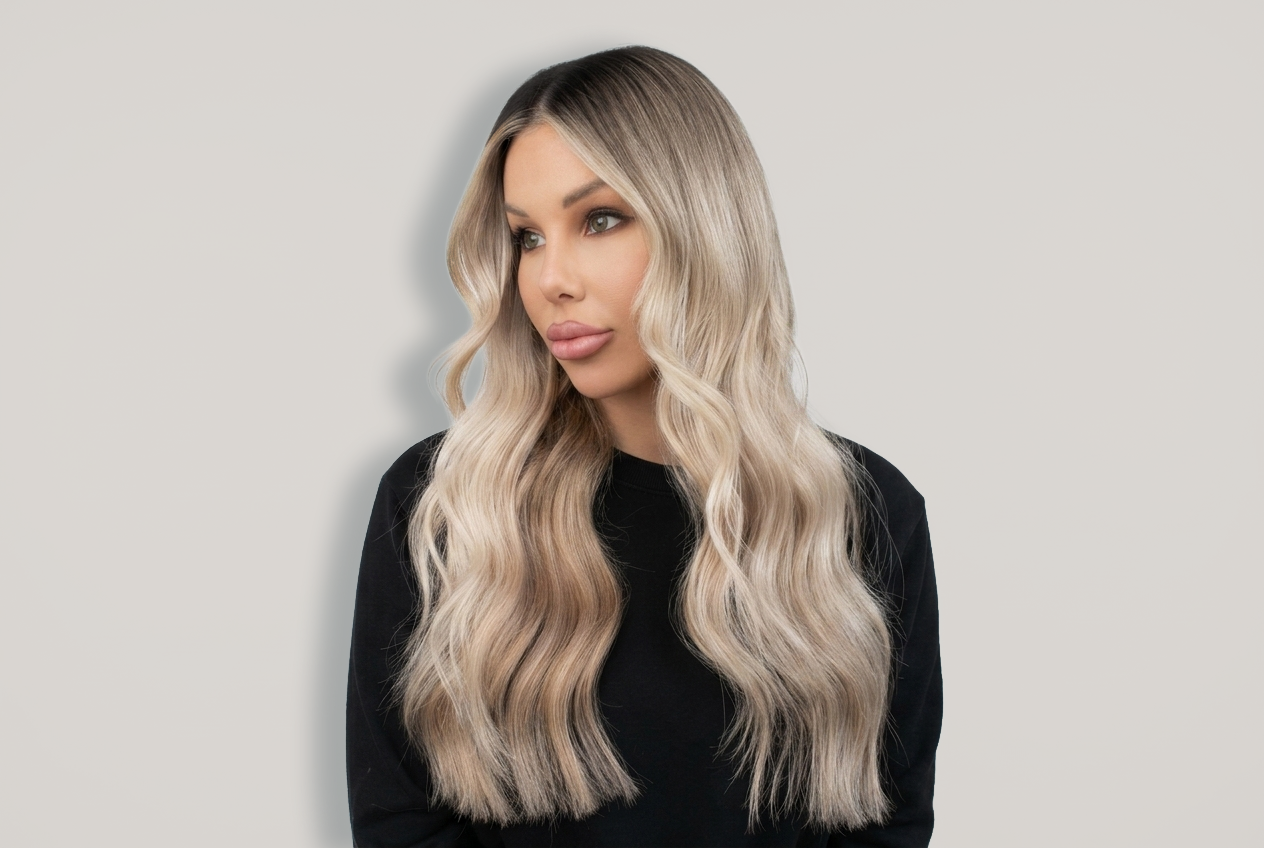 A woman with long, wavy blonde hair wearing a black top poses against a plain, light background.