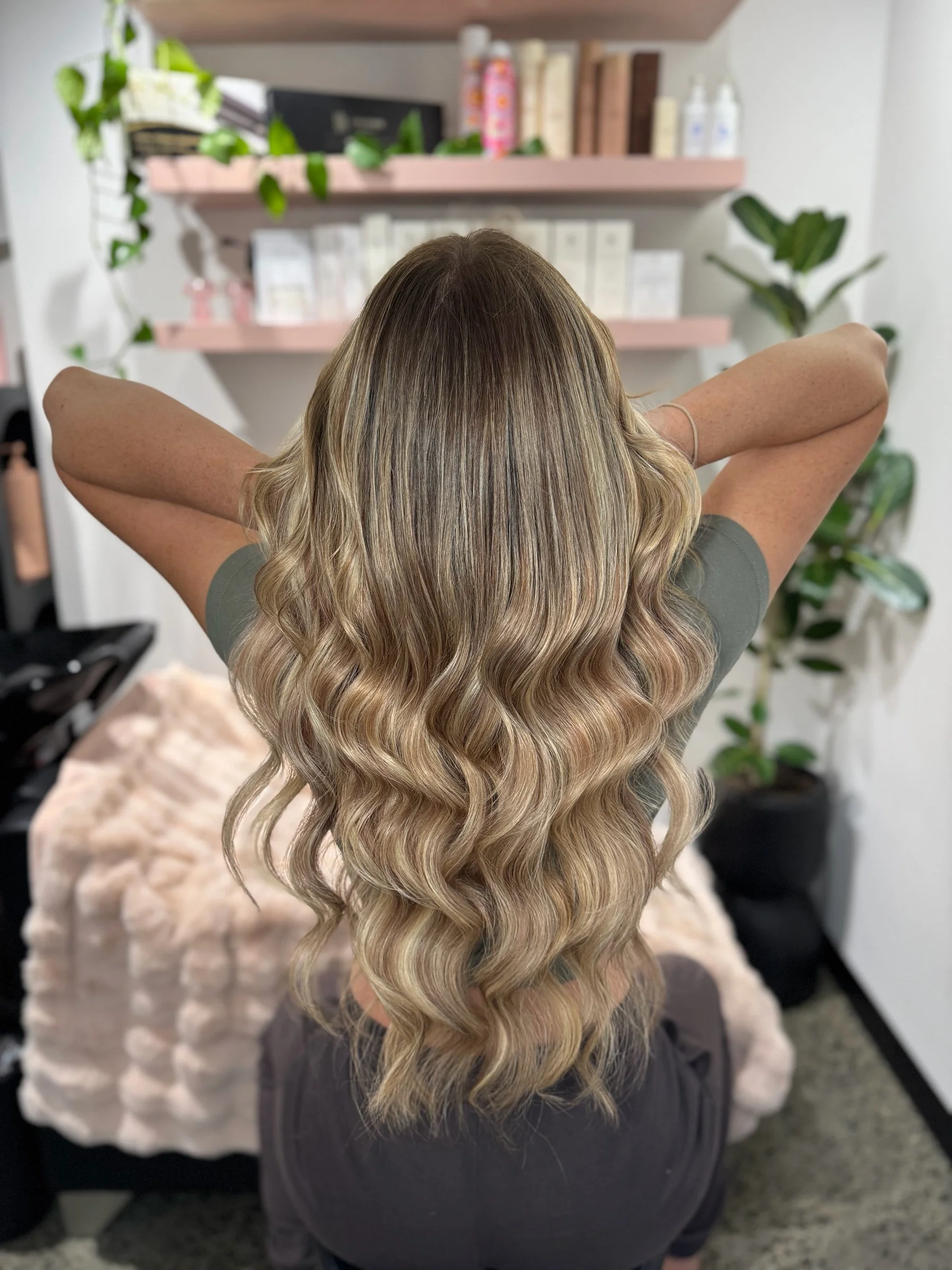 A woman sits with her arms behind her head, showing long, wavy blonde hair with darker roots in a salon or beauty studio, with shelves of hair products and plants in the background.
