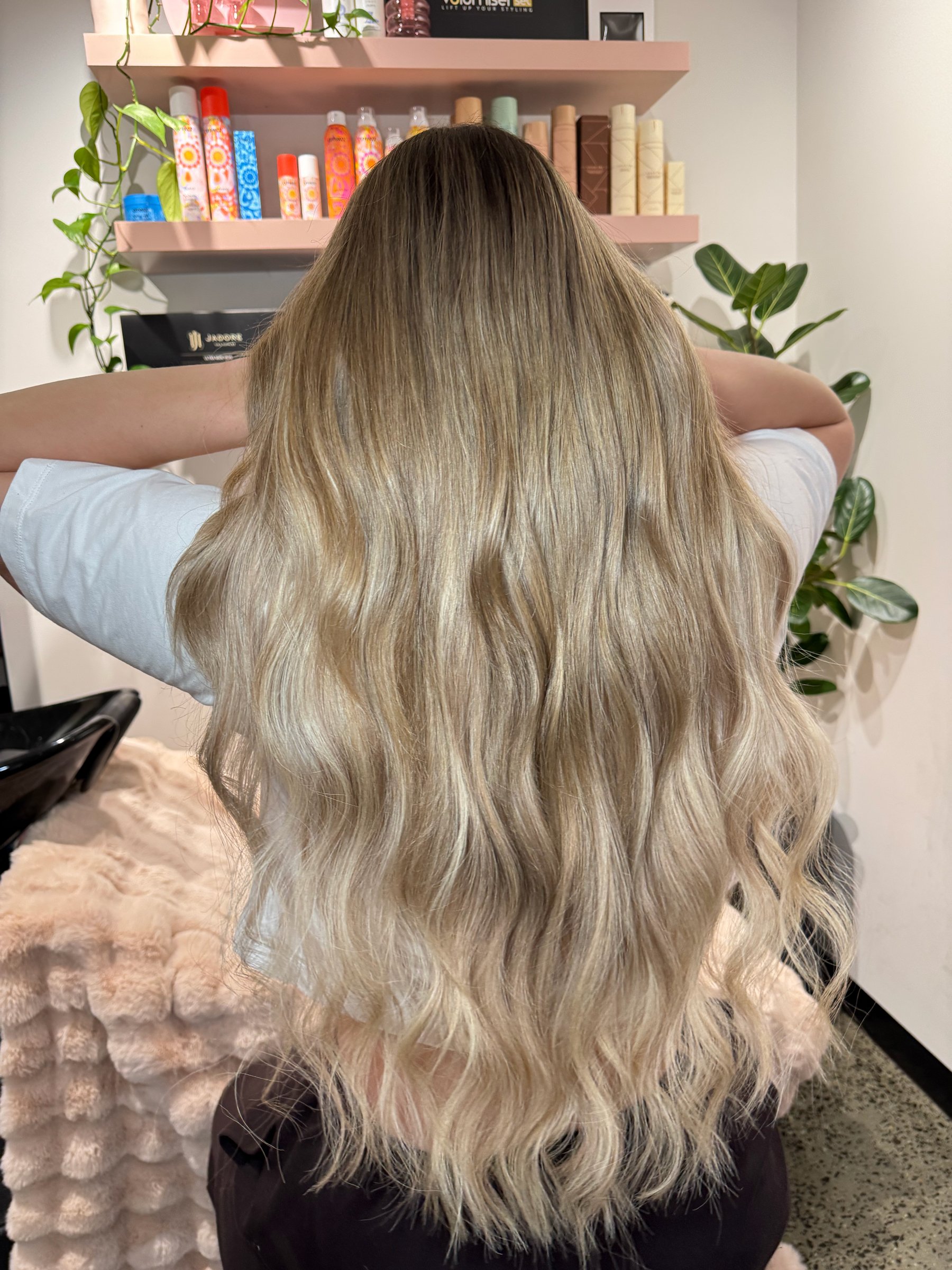 Back view of a woman with long, wavy blonde hair sitting in a salon. She has her arms raised behind her head. In the background, there are shelves with hair products and green plants.