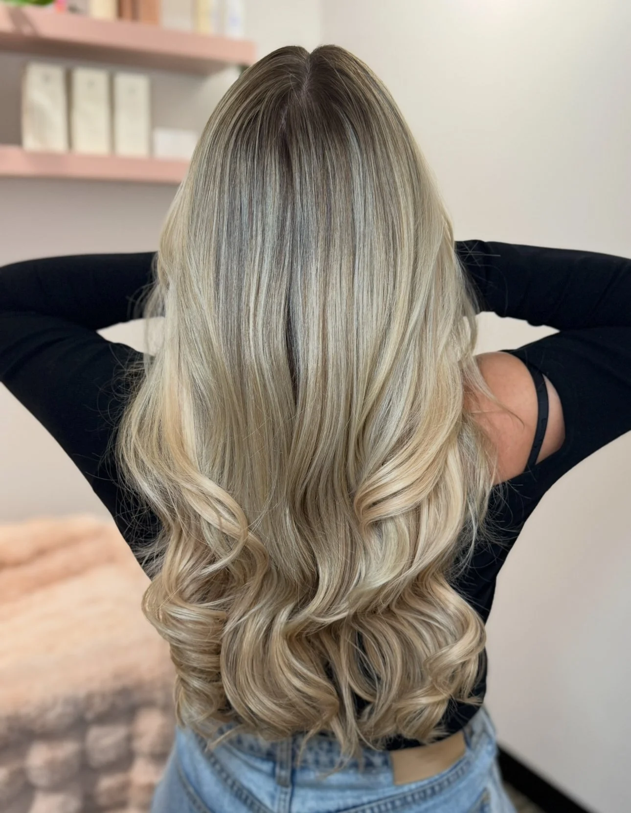 Woman with long, blonde, wavy hair, wearing a black top, standing indoors with her back to the camera.