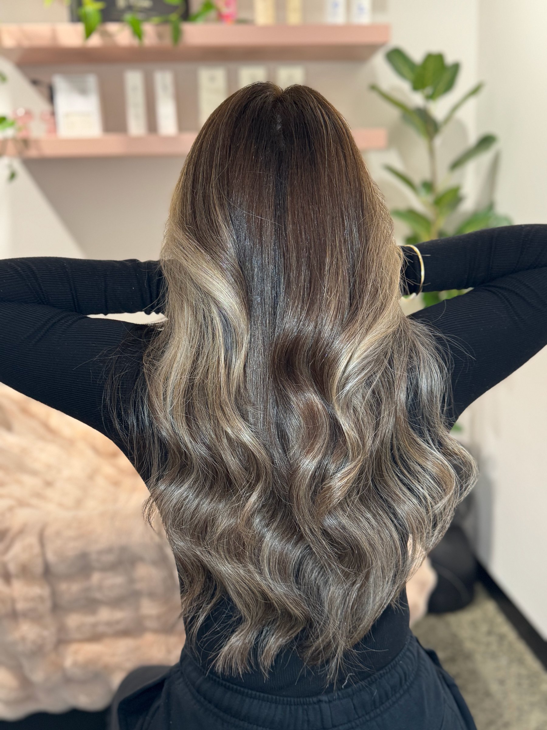 Back view of a person with long, wavy hair that is dark at the roots and fades to blonde. The person is wearing a black top and has arms behind the head. The background shows some indoor plants and blurred shelves.