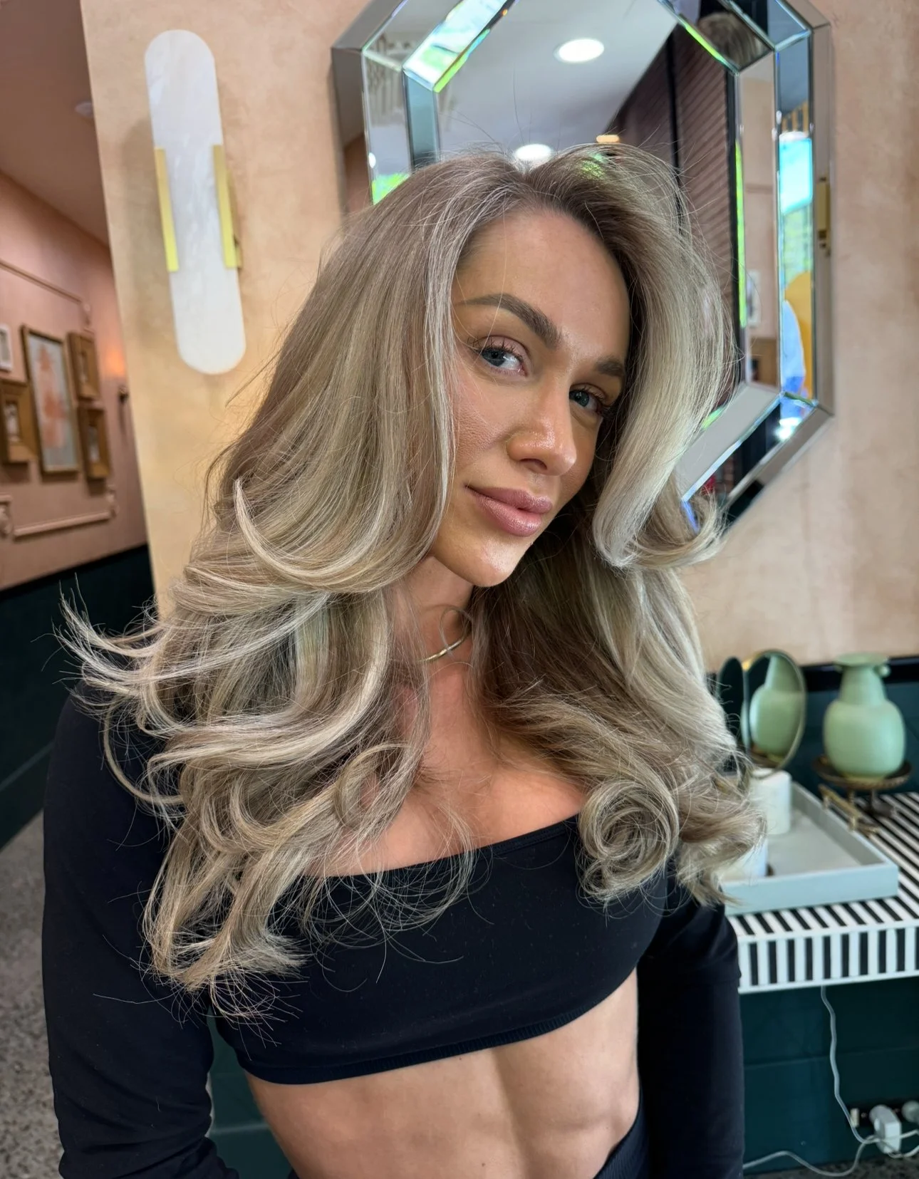 A woman with long, wavy blonde hair, wearing a black crop top, standing indoors with a mirror and decorative items in the background.