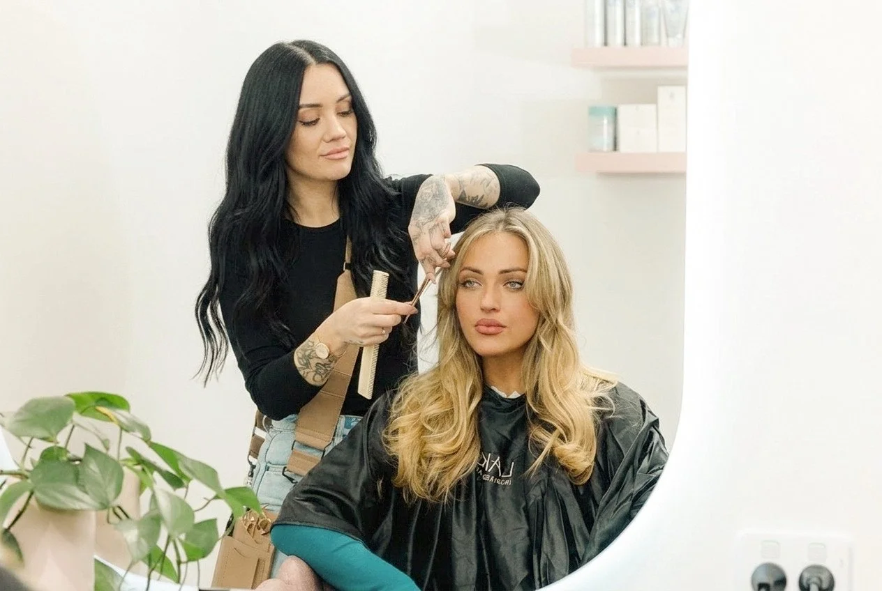 A hairstylist with dark hair and tattoos is styling a blonde woman's hair in a salon.