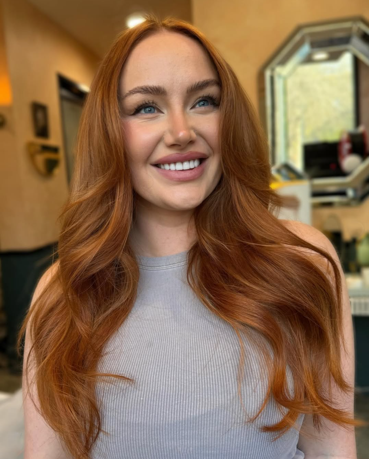 A young woman with long, wavy red hair, blue eyes, and a bright smile in a cozy indoor setting.