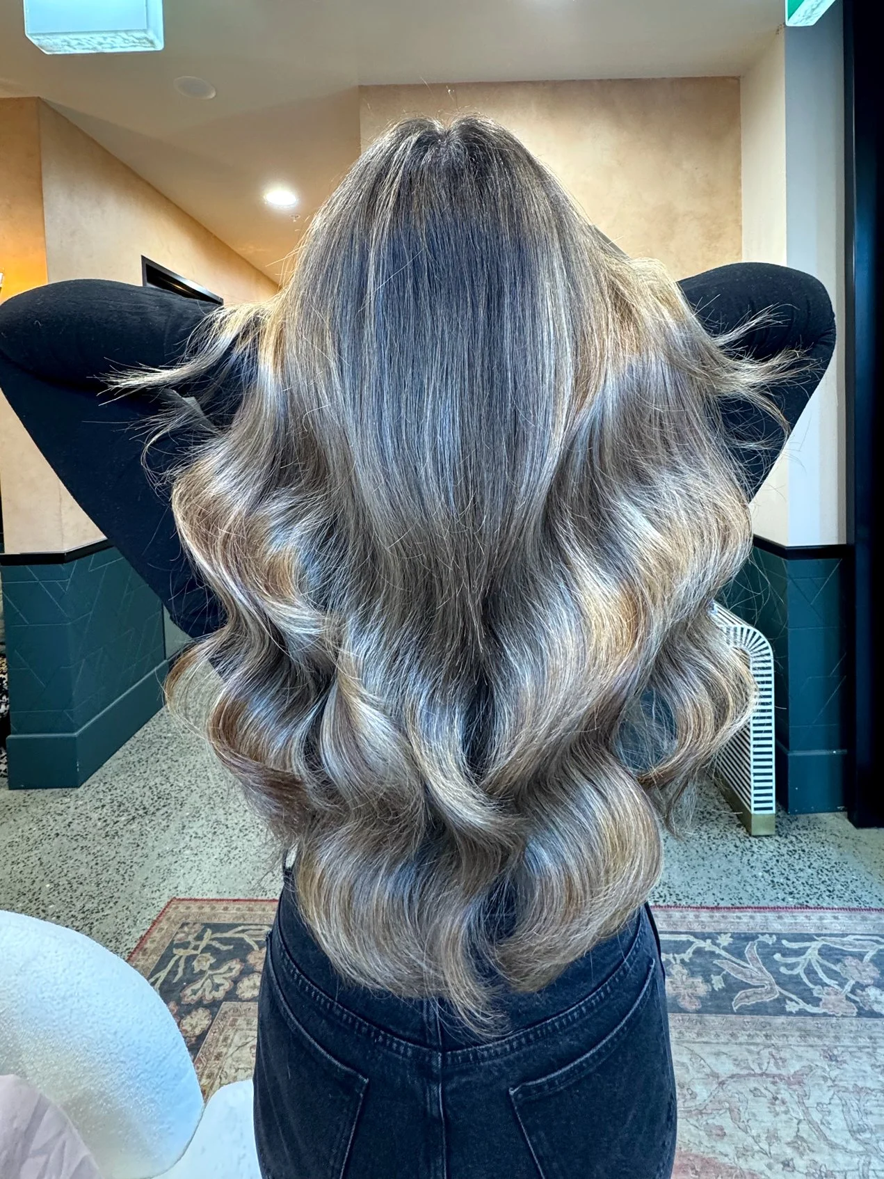 Back view of a woman with long, wavy, blonde hair standing in an indoor space, arms raised behind her head, wearing a black top and blue jeans.