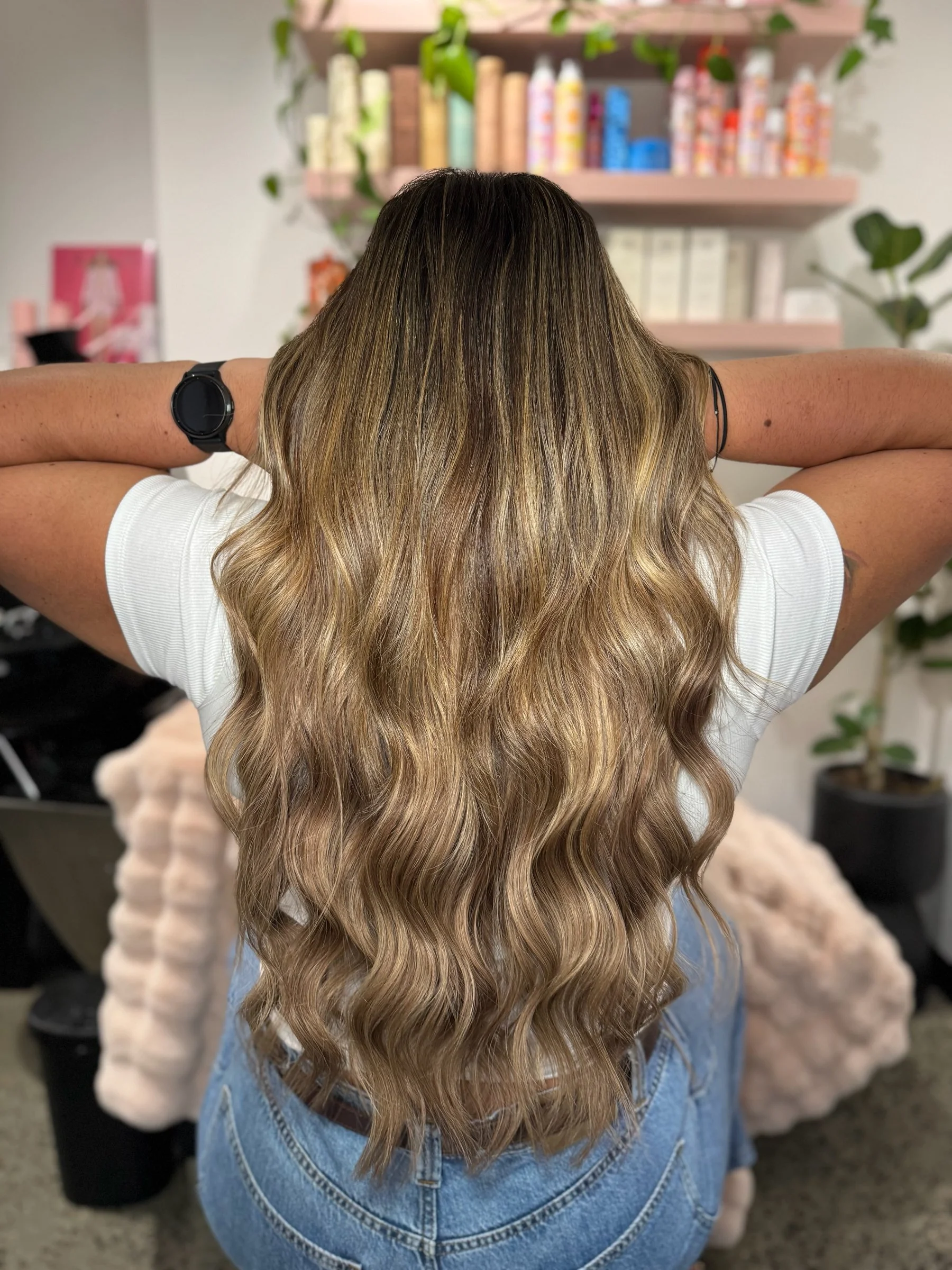 Back view of a woman with long, wavy, ombré hair in shades of dark blonde to light blonde, sitting on a beige furry blanket in a room with shelves of hair products in the background.