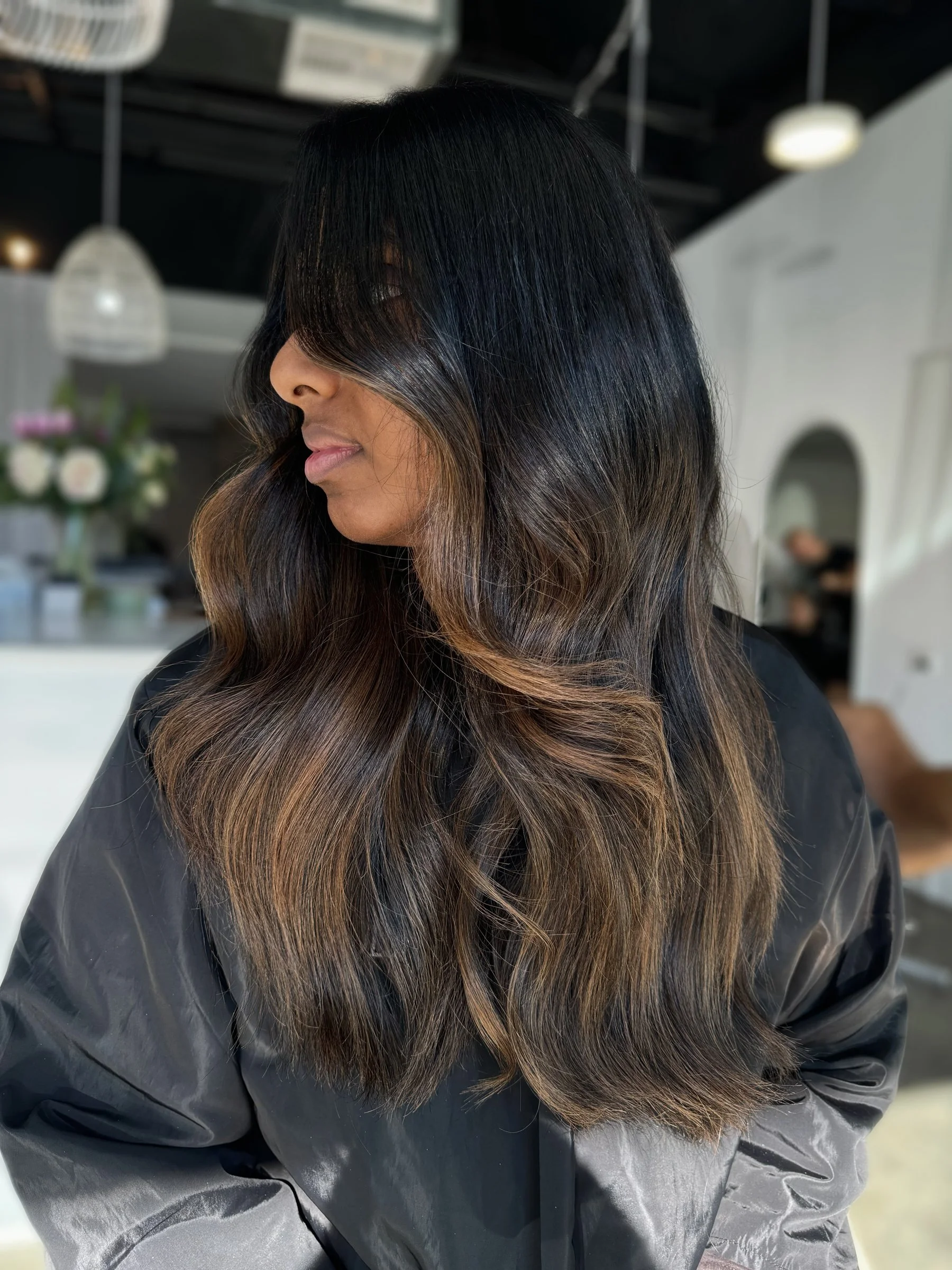 A woman with long, dark hair that transitions to light brown at the ends, styled in loose waves. She is in a hairstyle salon wearing a black cape, with a white wall and modern lighting fixtures in the background.
