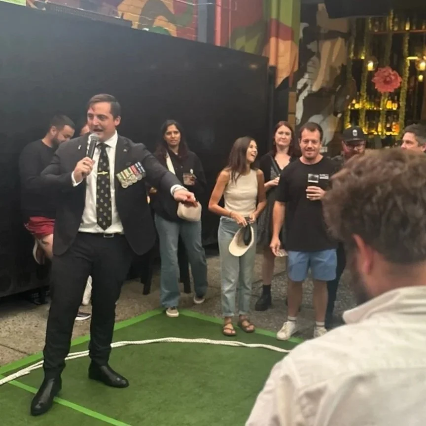 A man dressed in a suit with military medals is speaking into a microphone at an event, surrounded by a group of people listening and smiling, with a black backdrop and colorful wall art.