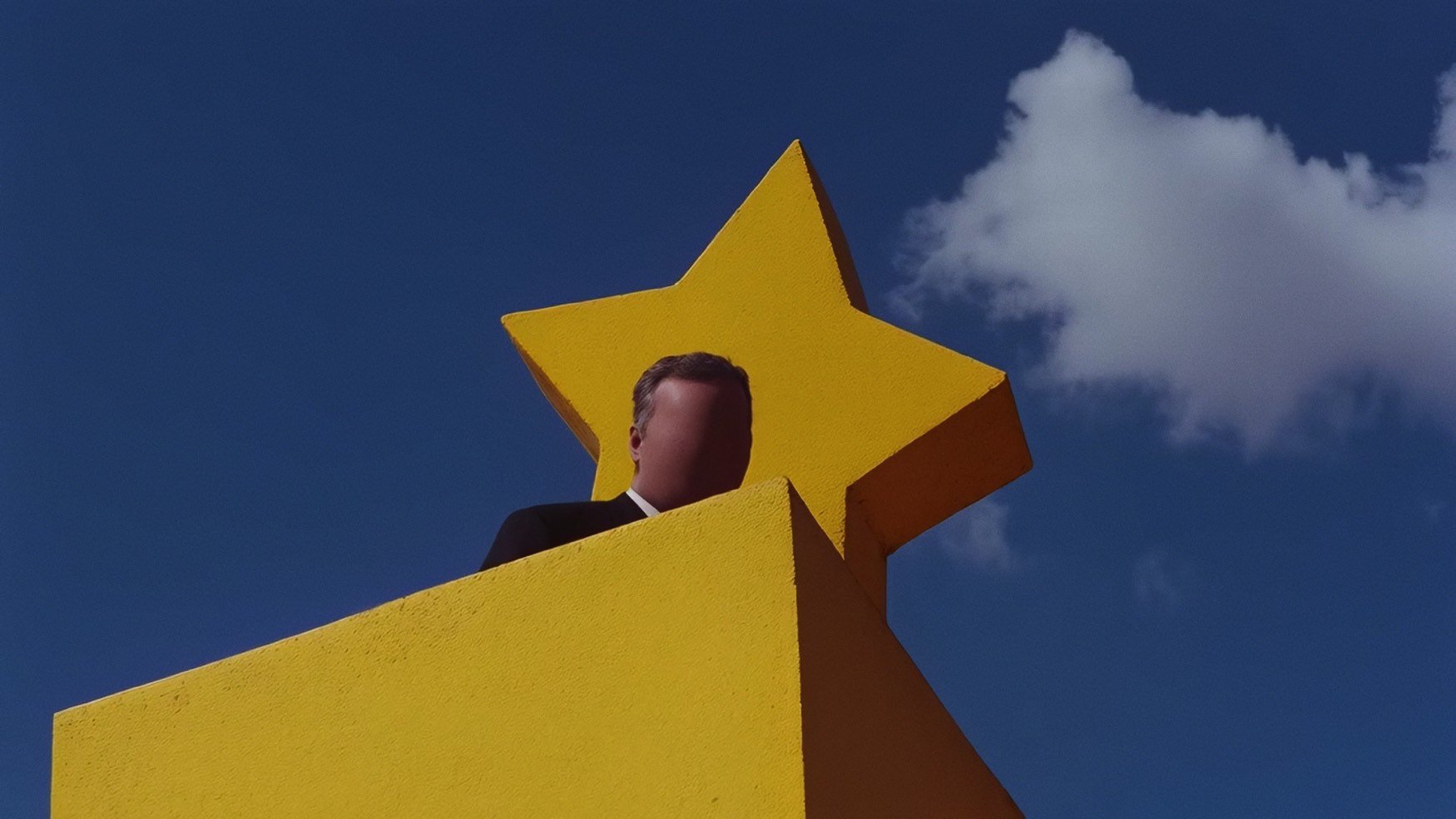 A person with a blurred face standing behind a large yellow star sculpture on a yellow platform against a blue sky with some clouds.