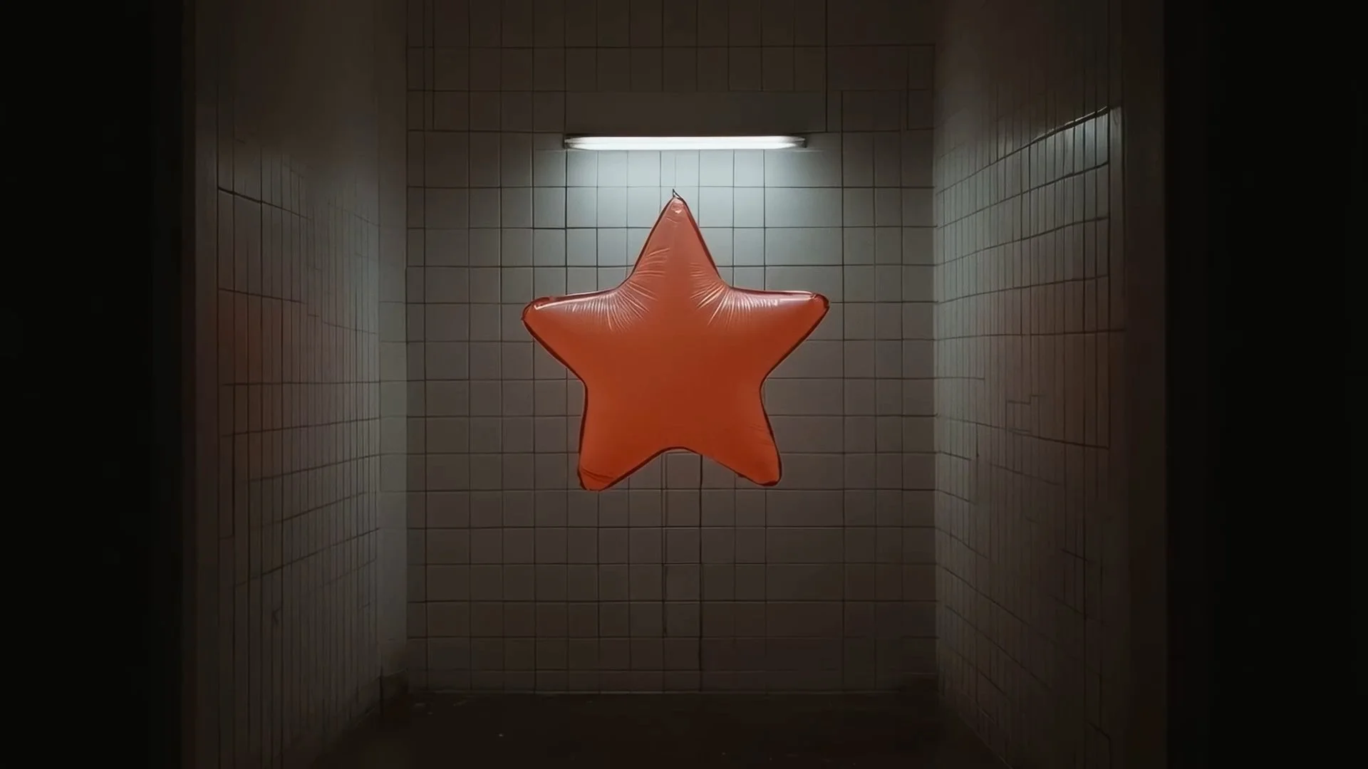 A large, orange, star-shaped balloon hanging in a tiled room with white walls and a fluorescent light above.