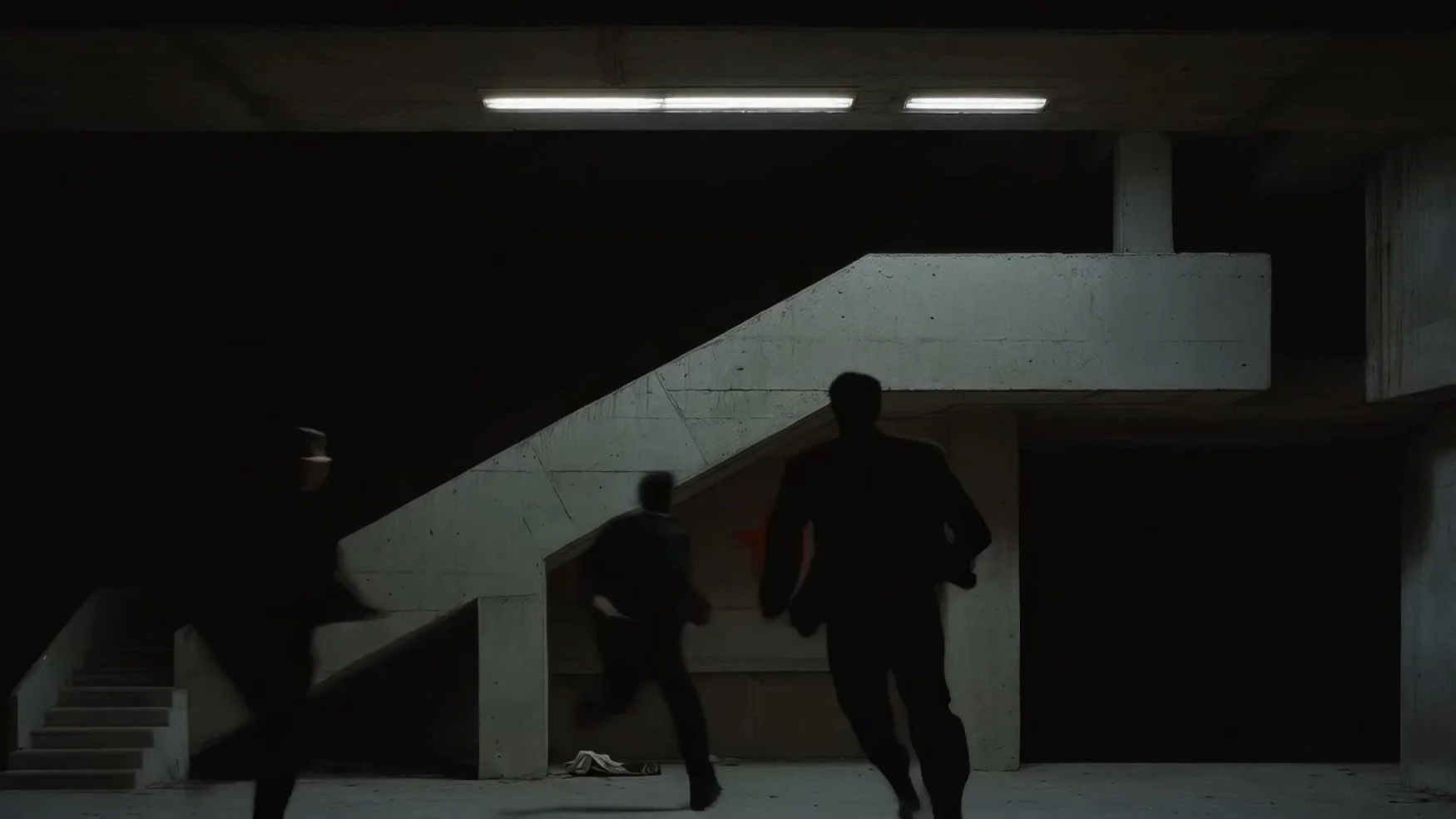Two people running under an overpass or parking garage with concrete stairs and structure, in a dimly lit indoor setting.