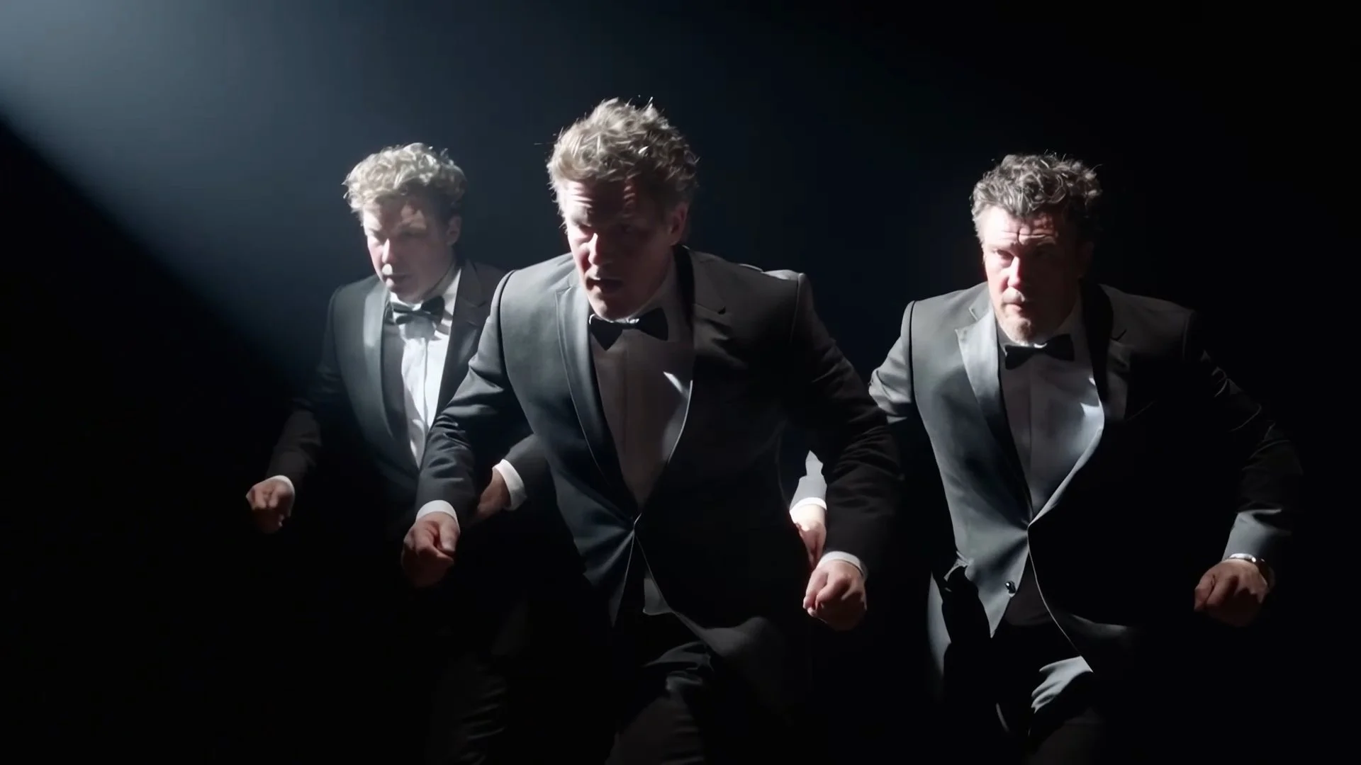 Three men in tuxedos perform a synchronized dance in a dark setting with a spotlight.